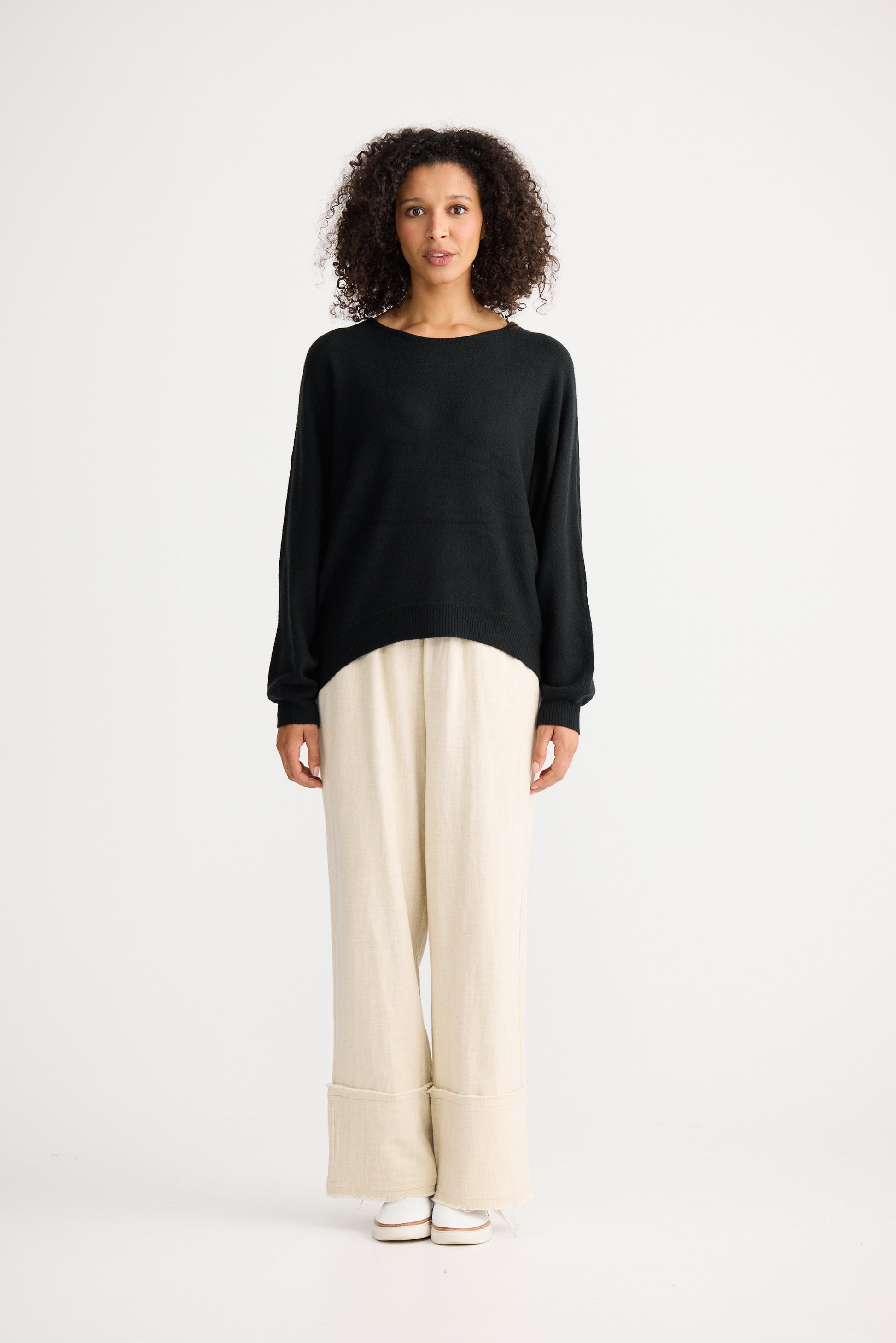 Woman wearing a Shanty Corporation black sweater and beige pants on a white background