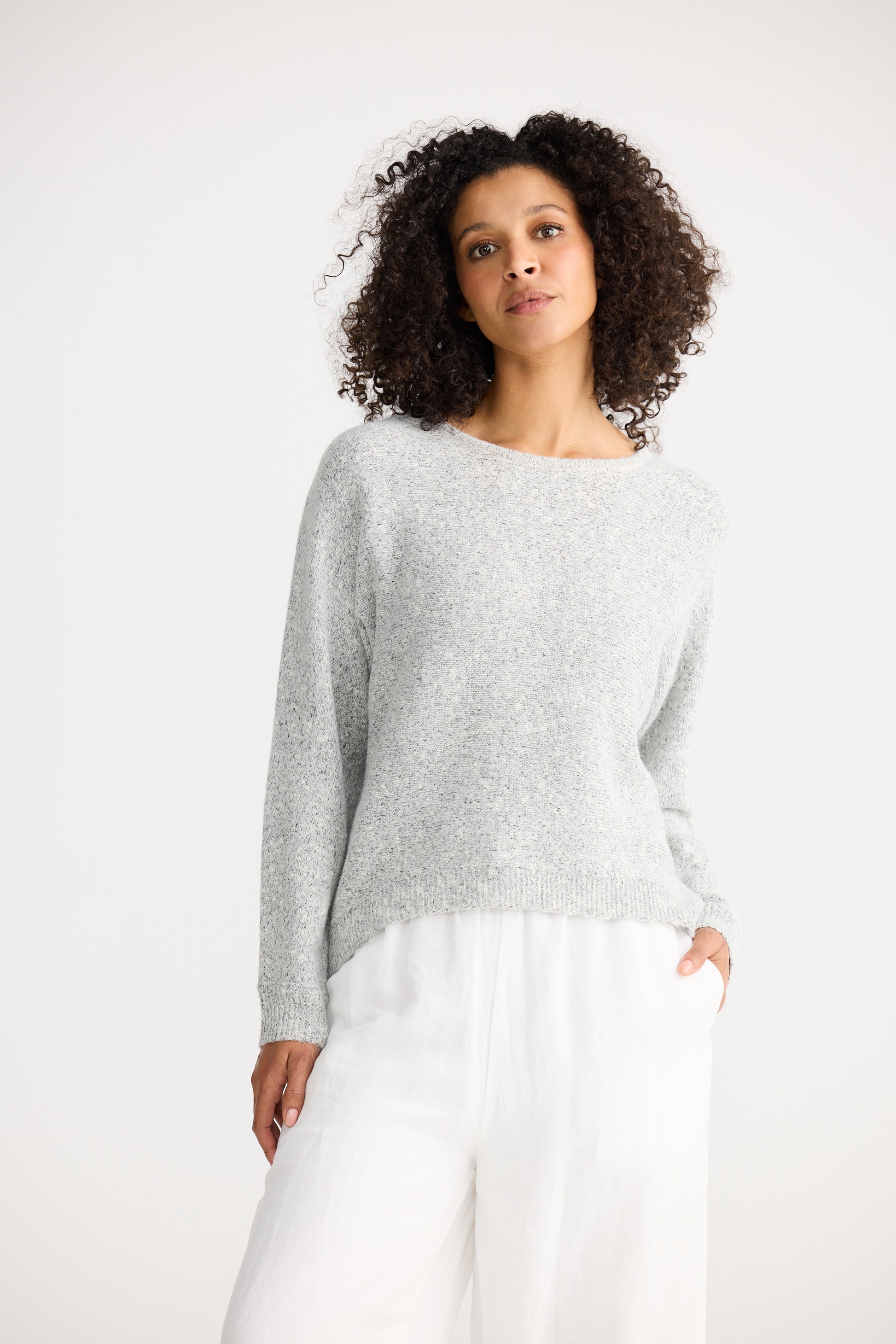 Woman wearing a Shanty Corporation light grey sweater and white pants on a white background