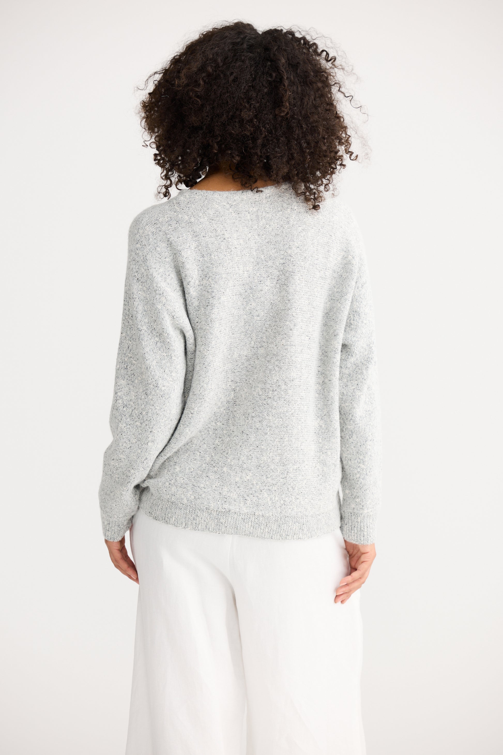 Woman wearing a Shanty Corporation light grey sweater and white pants on a plain background