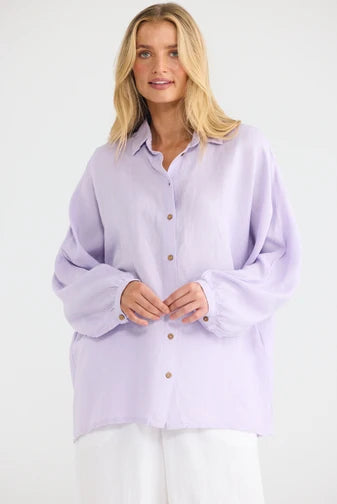 A woman wearing a Shanty Corporation light lavender oversized button-up shirt with long sleeves and white pants, standing against a plain white studio background.