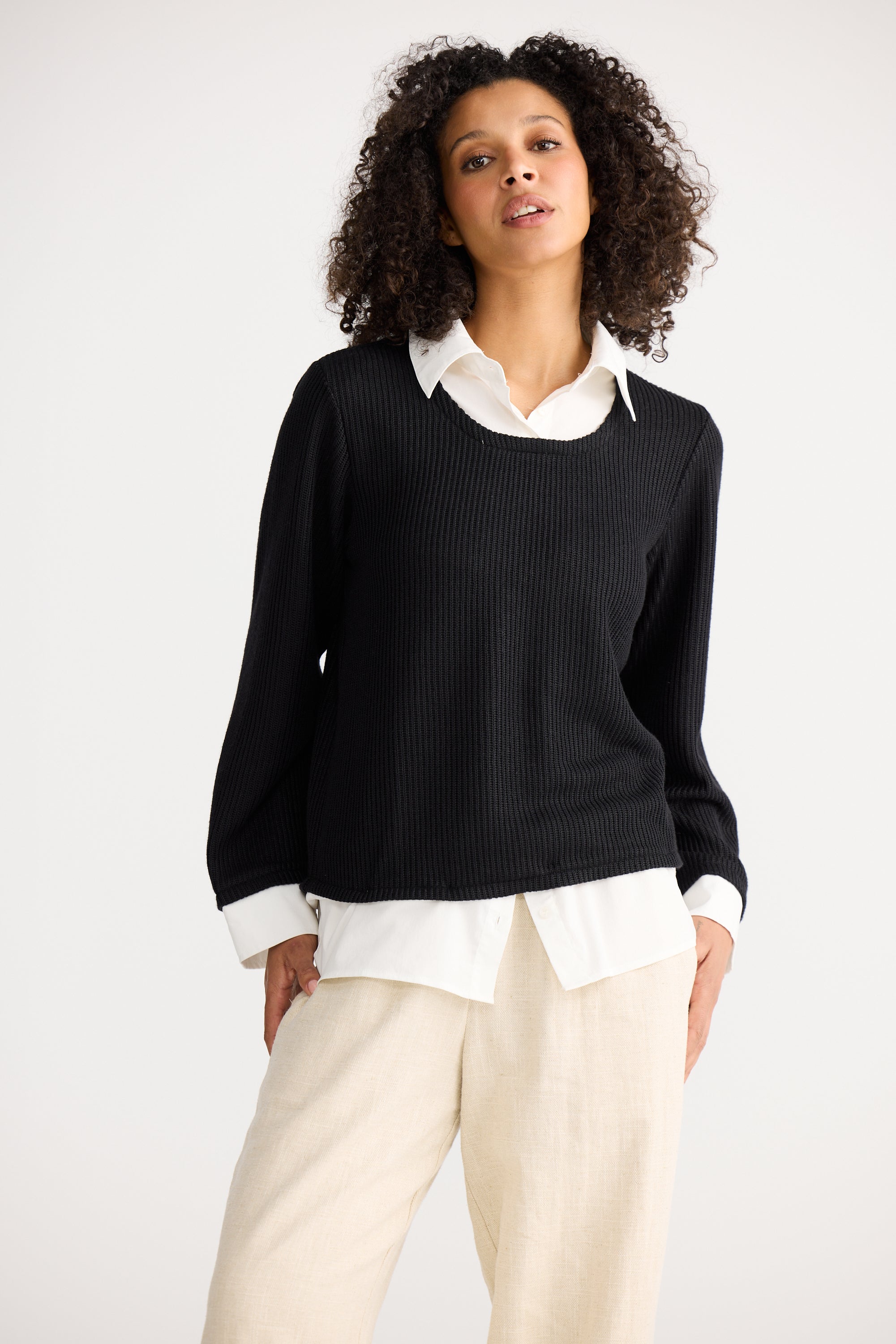 Image of Woman wearing a Shanty Corporation black sweater over a white shirt with beige pants on a white background