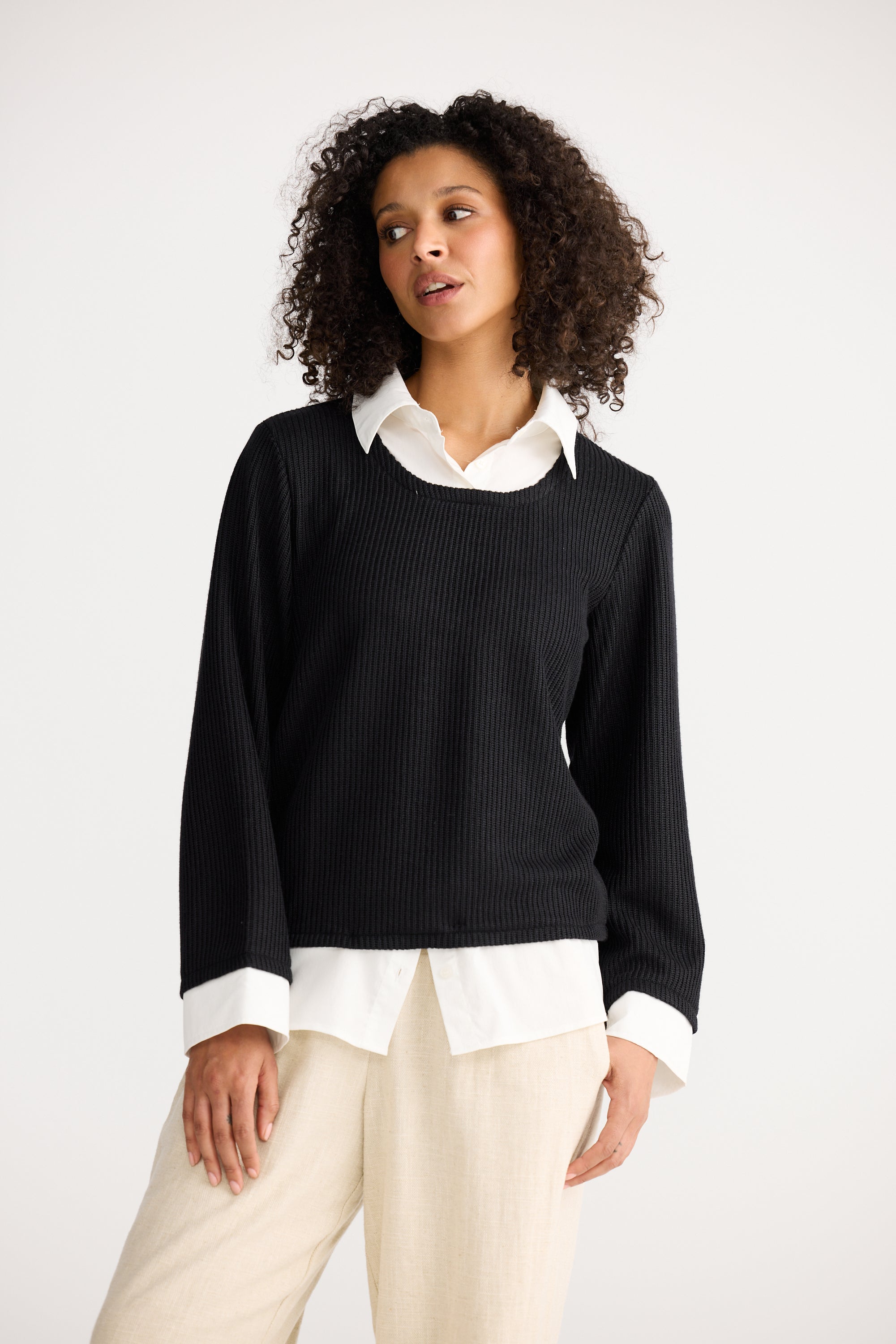 Image of Woman wearing a Shanty Corporation black sweater over a white shirt with beige pants on a white background