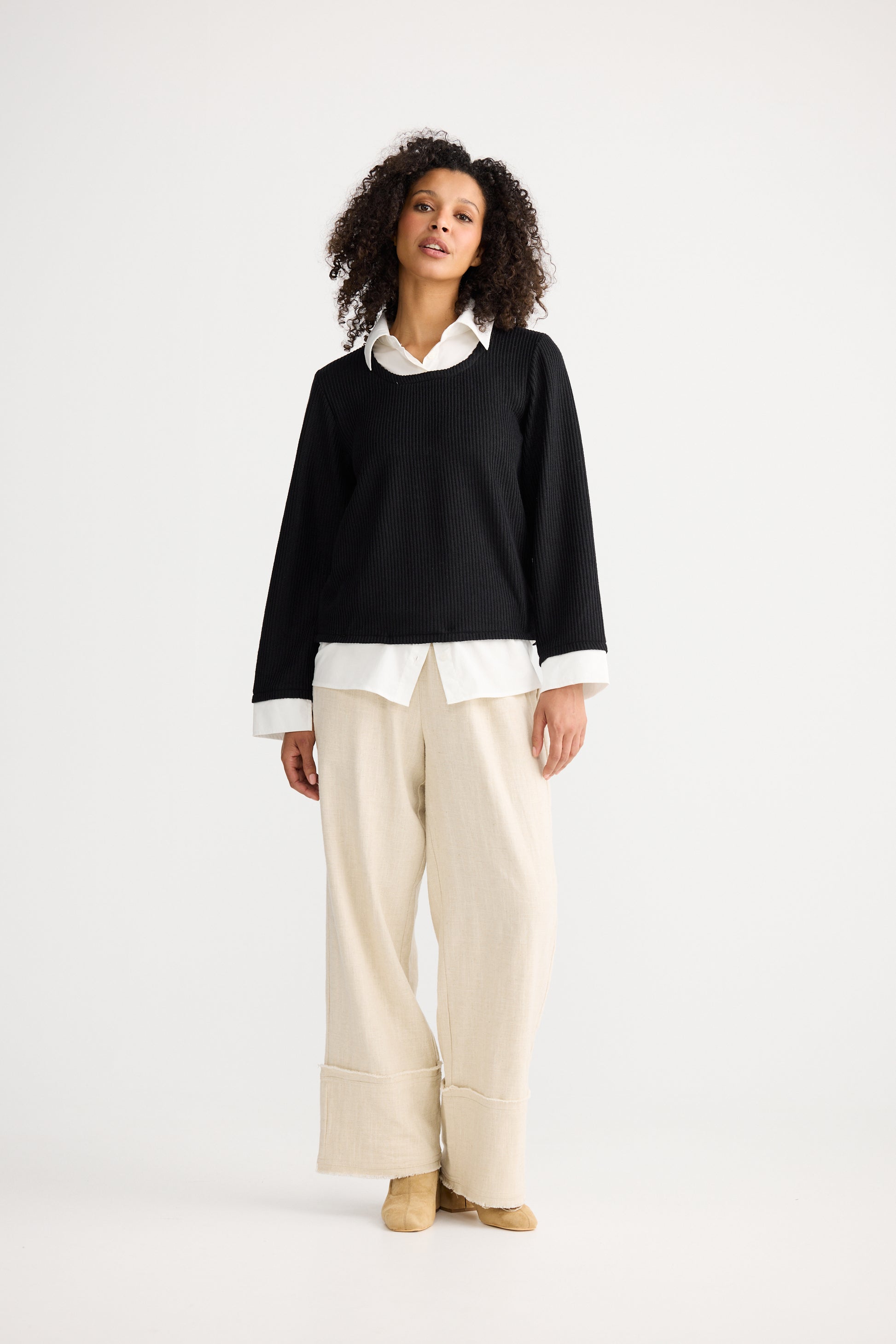 Woman wearing a Shanty Corporation black sweater over a white shirt with beige pants on a white background
