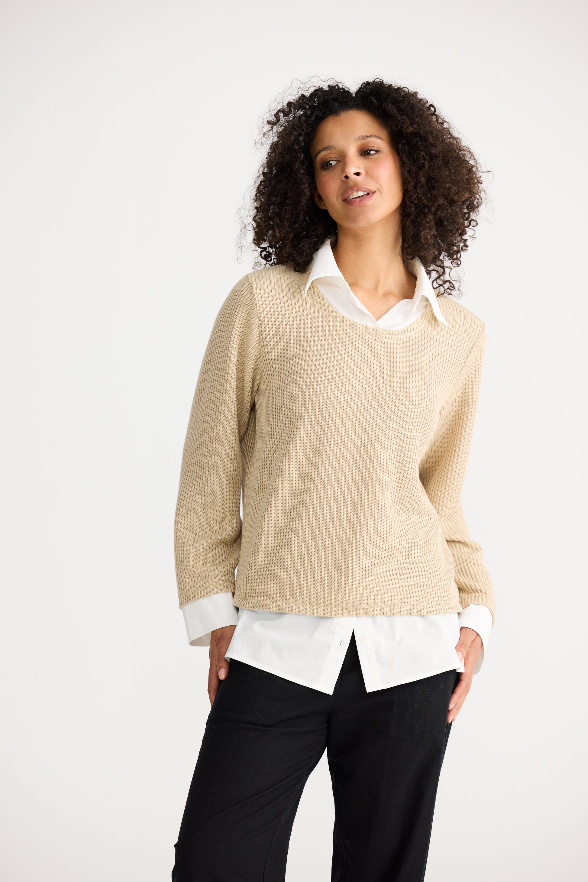 Woman wearing a Shanty Corporation beige sweater over a white shirt with black pants on a white background
