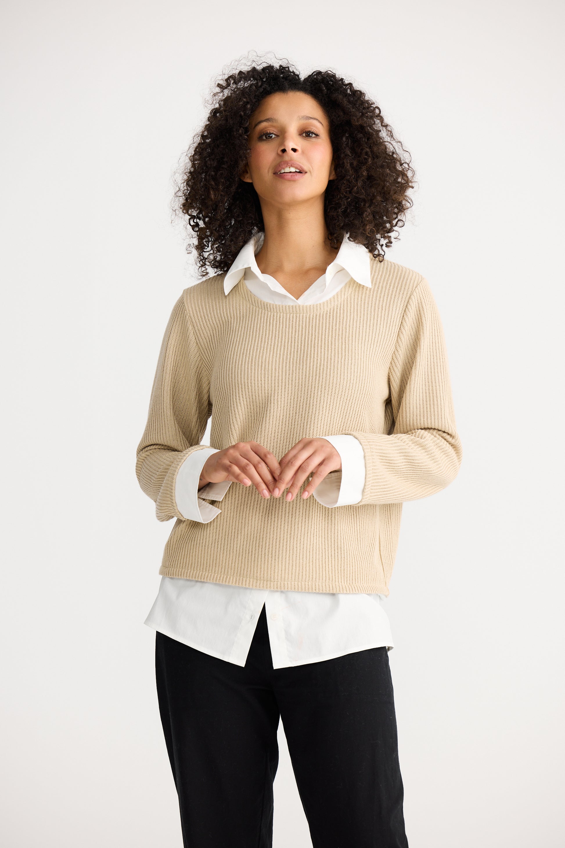 Woman wearing a Shanty Corporation beige sweater over a white shirt with black pants on a white background