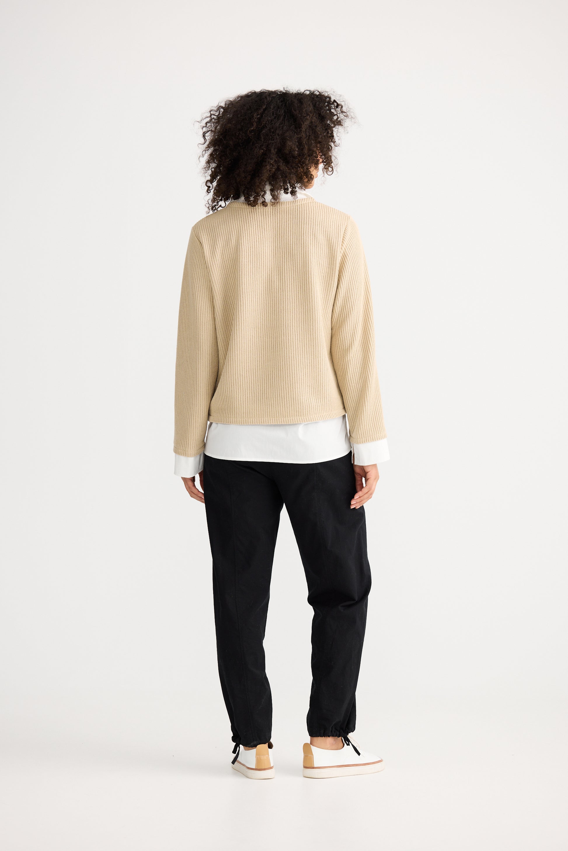 Woman wearing a beige sweater over a white shirt with black pants on a white background