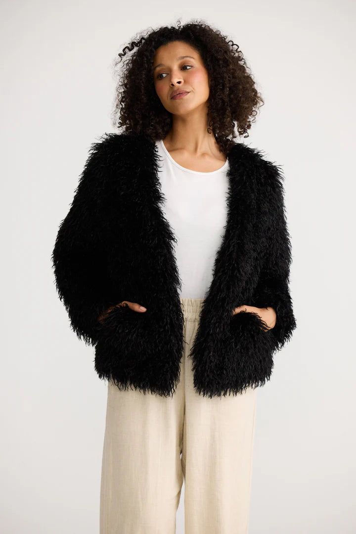 Image of Woman wearing a Shanty Corporation black fluffy jacket over a white top and beige pants on a white background