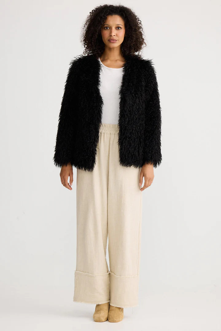 Woman wearing a Shanty Corporation black fluffy jacket, white top, and beige pants on a white background