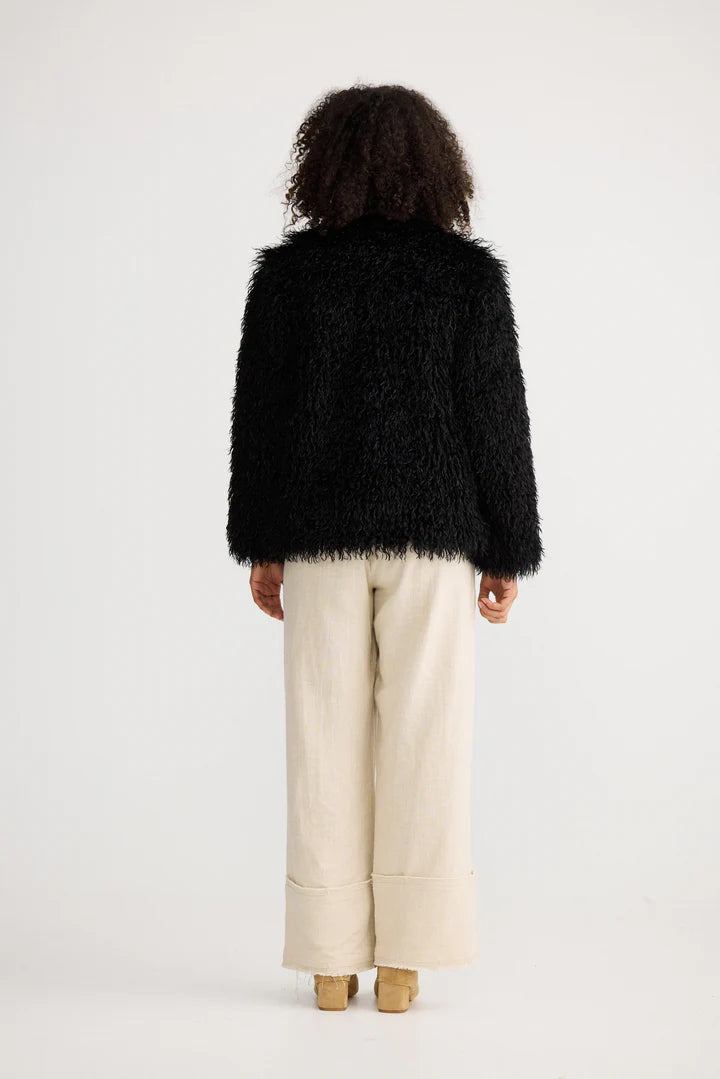 Woman wearing a Shanty Corporation black fluffy jacket and beige pants on a white background