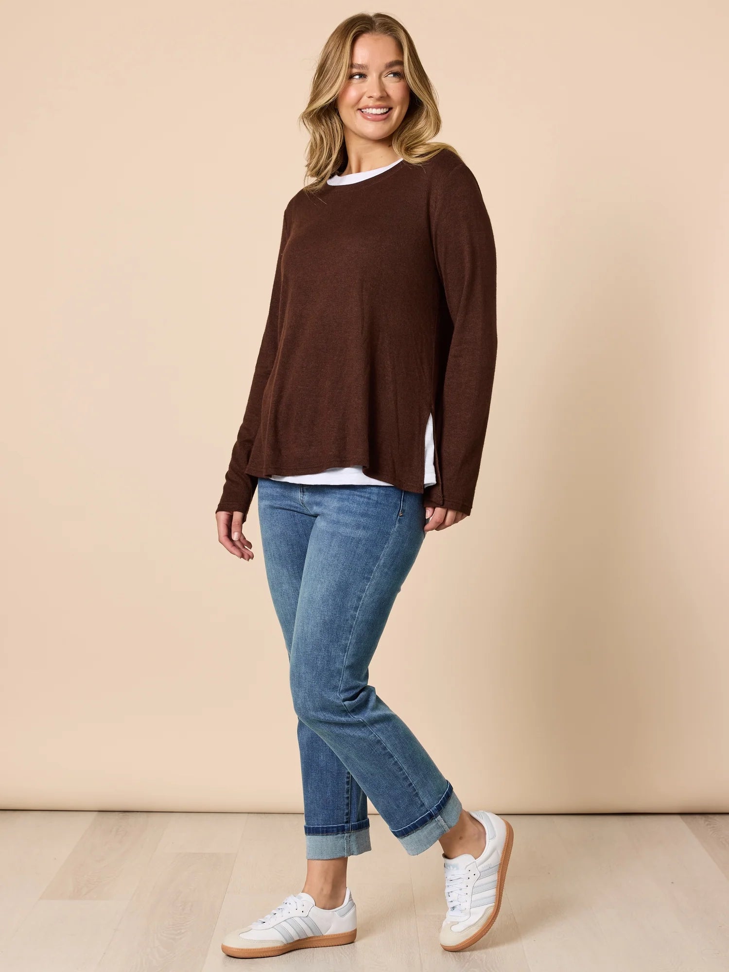 Woman wearing a Threadz brown sweater, white shirt, blue jeans, and white sneakers on a beige background