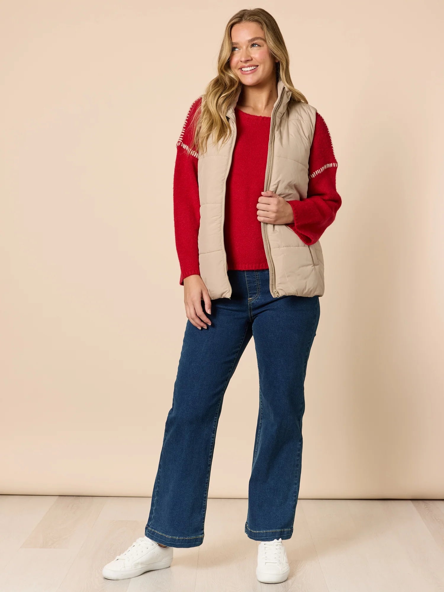 Woman wearing a red sweater, beige vest, and Threadz blue jeans against a beige background