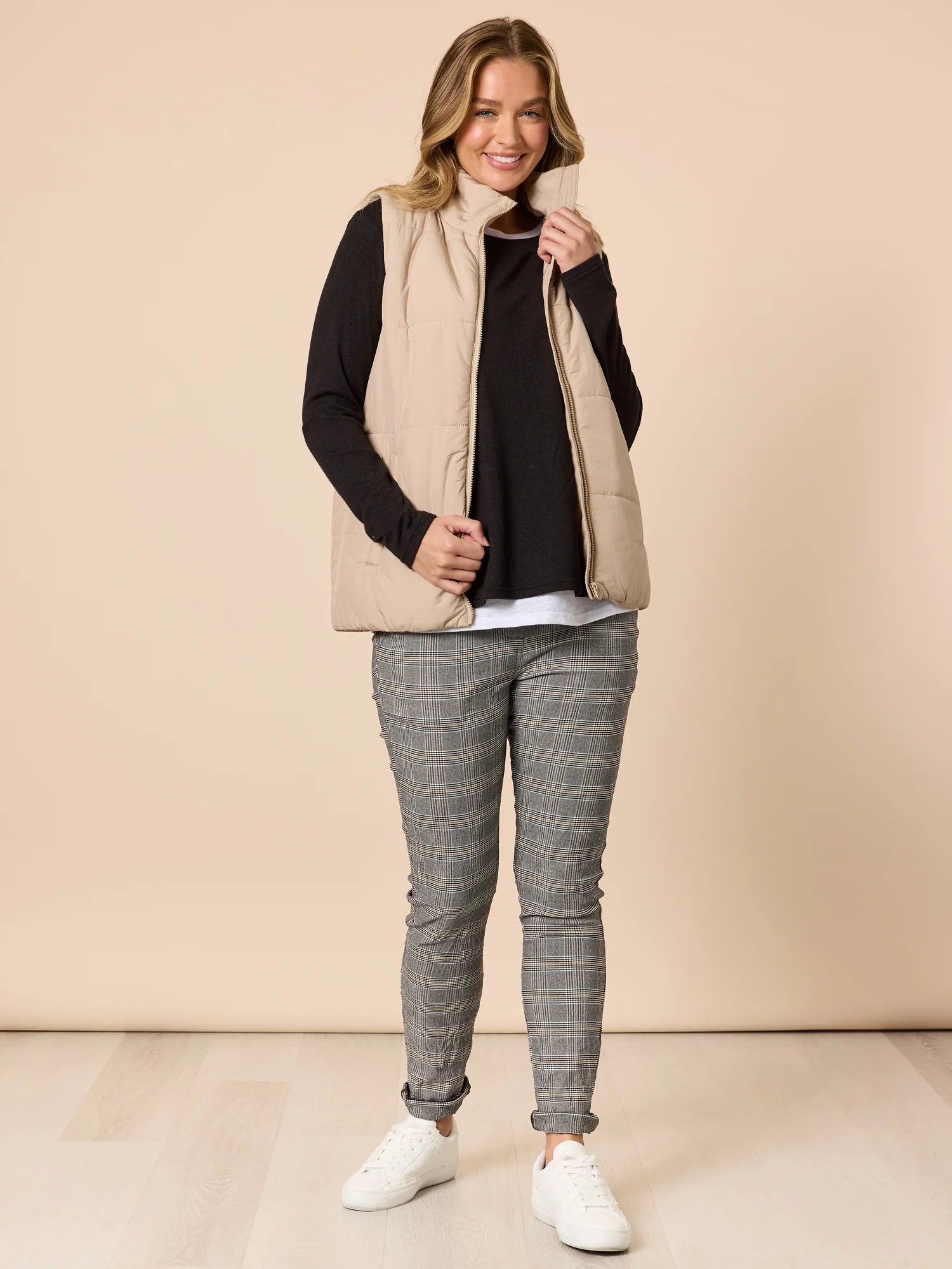 Woman wearing a beige vest over a Threadz black long-sleeve shirt, gray checkered pants, and white sneakers on a beige background.
