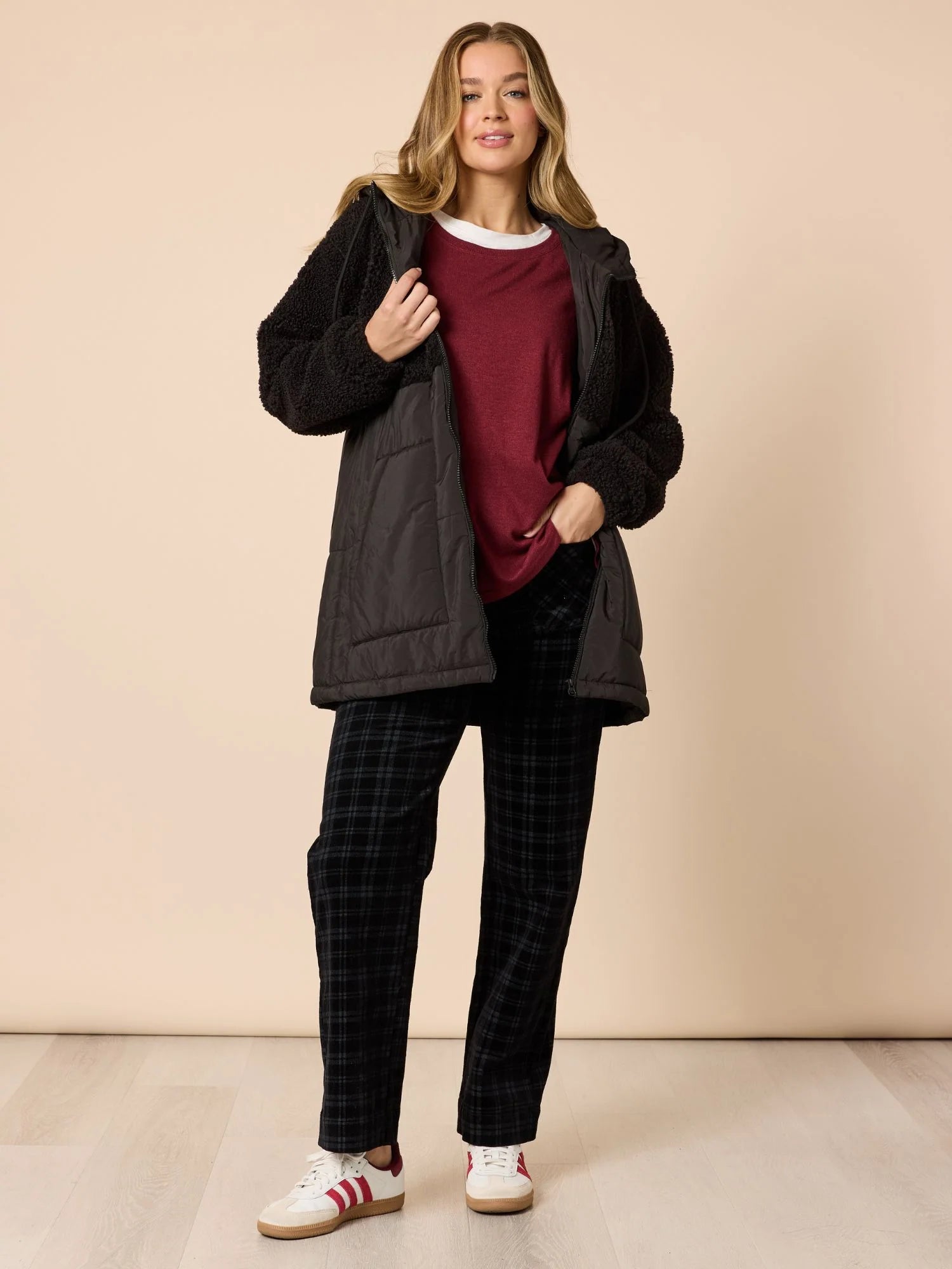 Woman wearing a black jacket, Threadz red shirt, plaid pants, and white sneakers on a beige background