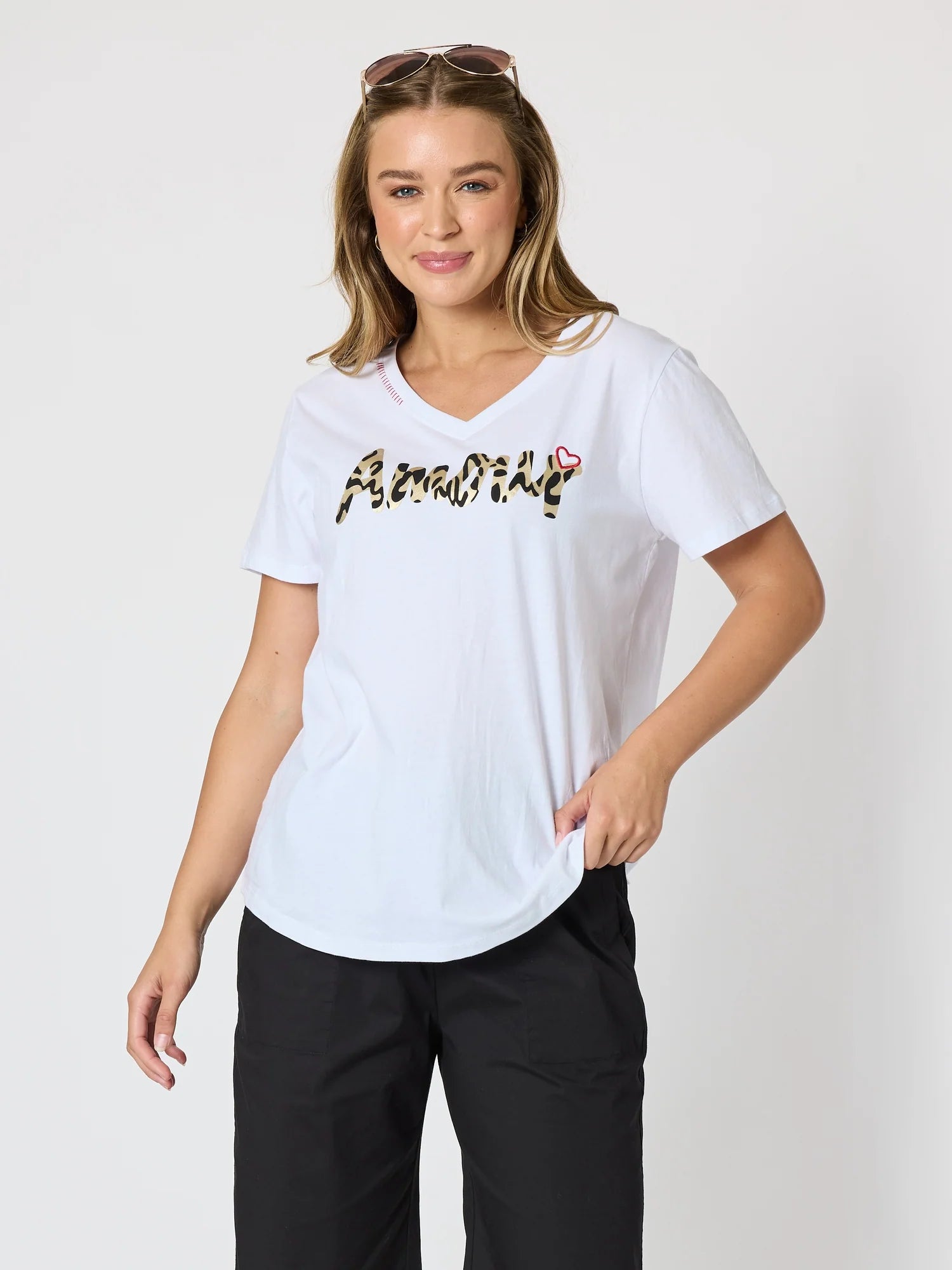 Image of A woman modeling a Threadz white short-sleeve V-neck T-shirt with a black and gold leopard-print graphic reading 'Amour' across the chest, paired with black pants, against a light gray studio background.