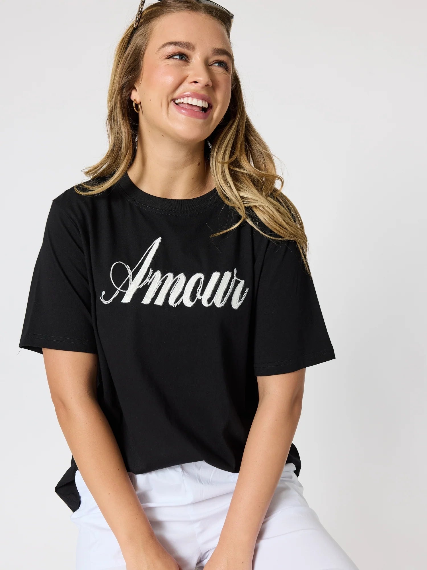 Image of A woman wearing a Threadz black short-sleeve crop top with 'Amour' printed in white script across the chest, paired with white pants, photographed against a light gray studio background.