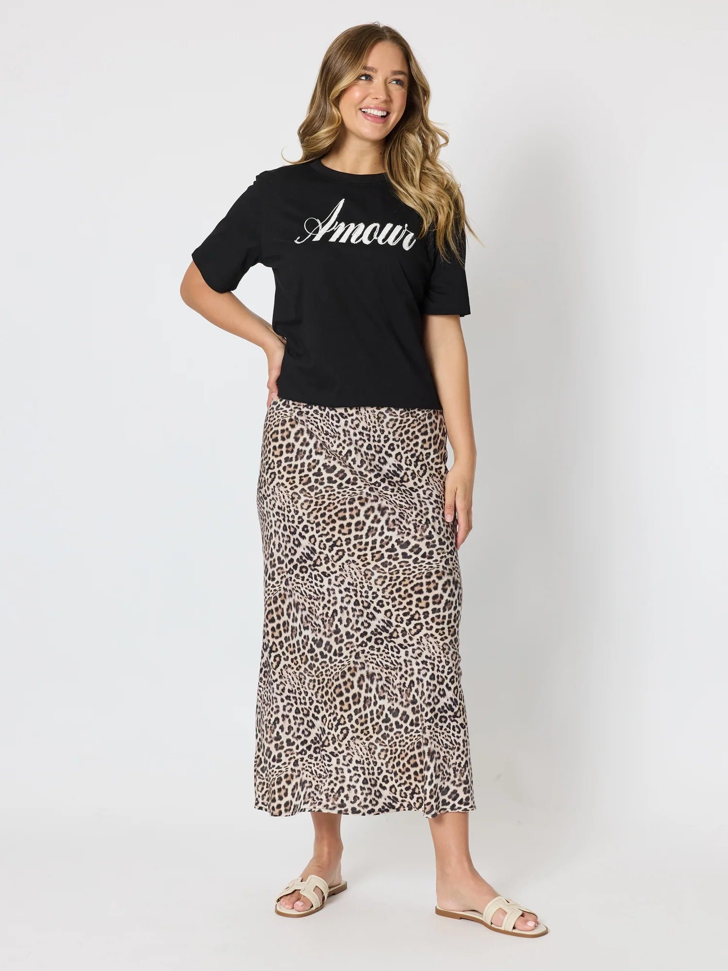 Image of A woman modeling a Threadz black short-sleeve t-shirt with 'Amour' printed in white script across the chest, paired with a leopard-print midi skirt and beige slide sandals, against a light gray studio background.