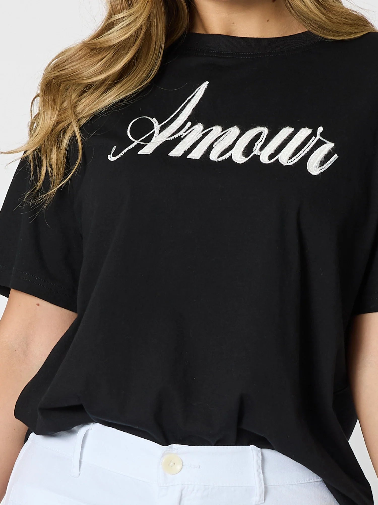 A close-up of a woman wearing a Threadz black short-sleeve T-shirt with the word 'Amour' printed across the chest in a white cursive script, paired with white pants.