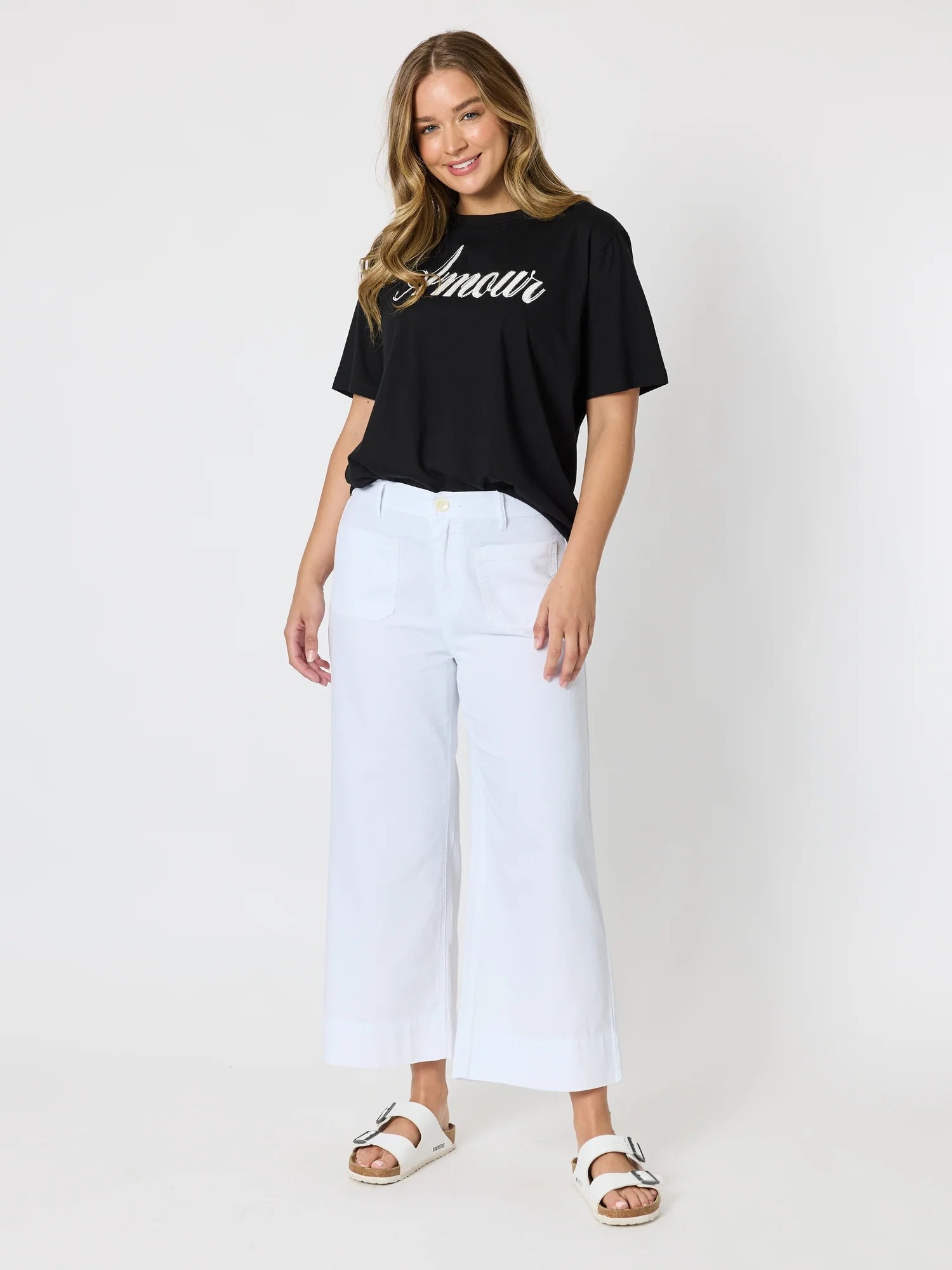Image of A woman modeling a black short-sleeve t-shirt with 'Amour' written in white script across the chest, paired with white cropped wide-leg pants and white slide sandals, against a plain light gray studio background.