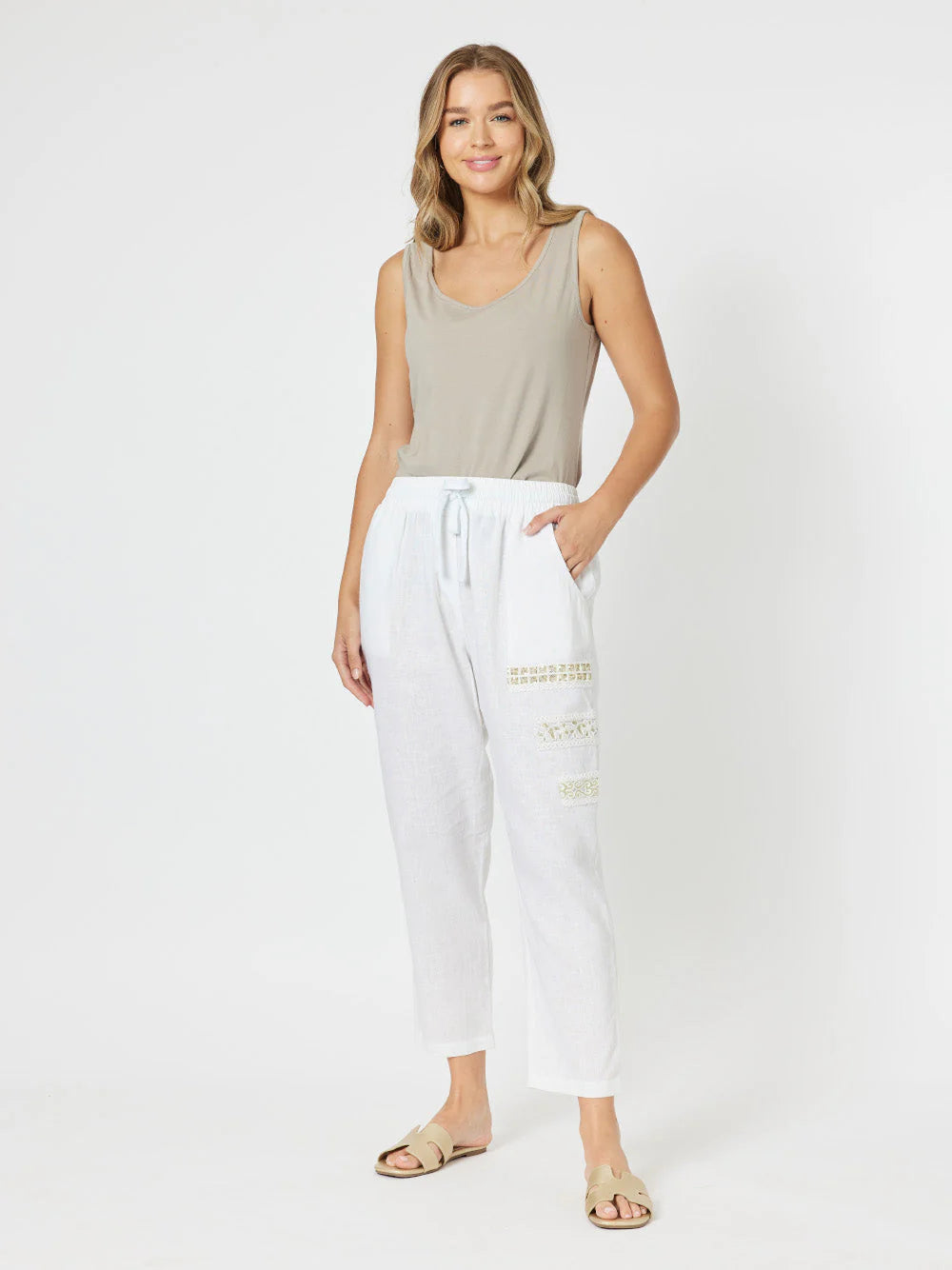 Image of A woman modeling white cropped drawstring pants with subtle gold text graphics on the lower legs, paired with a Threadz beige tank top and beige sandals, against a plain light gray studio background.