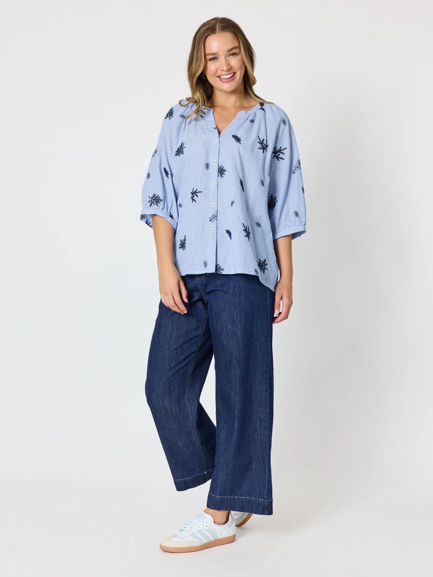 Image of A woman modeling a Threadz light blue short-sleeve button-up shirt with a dark navy palm tree print, paired with dark blue wide-leg jeans and white sneakers, against a plain light gray studio background.