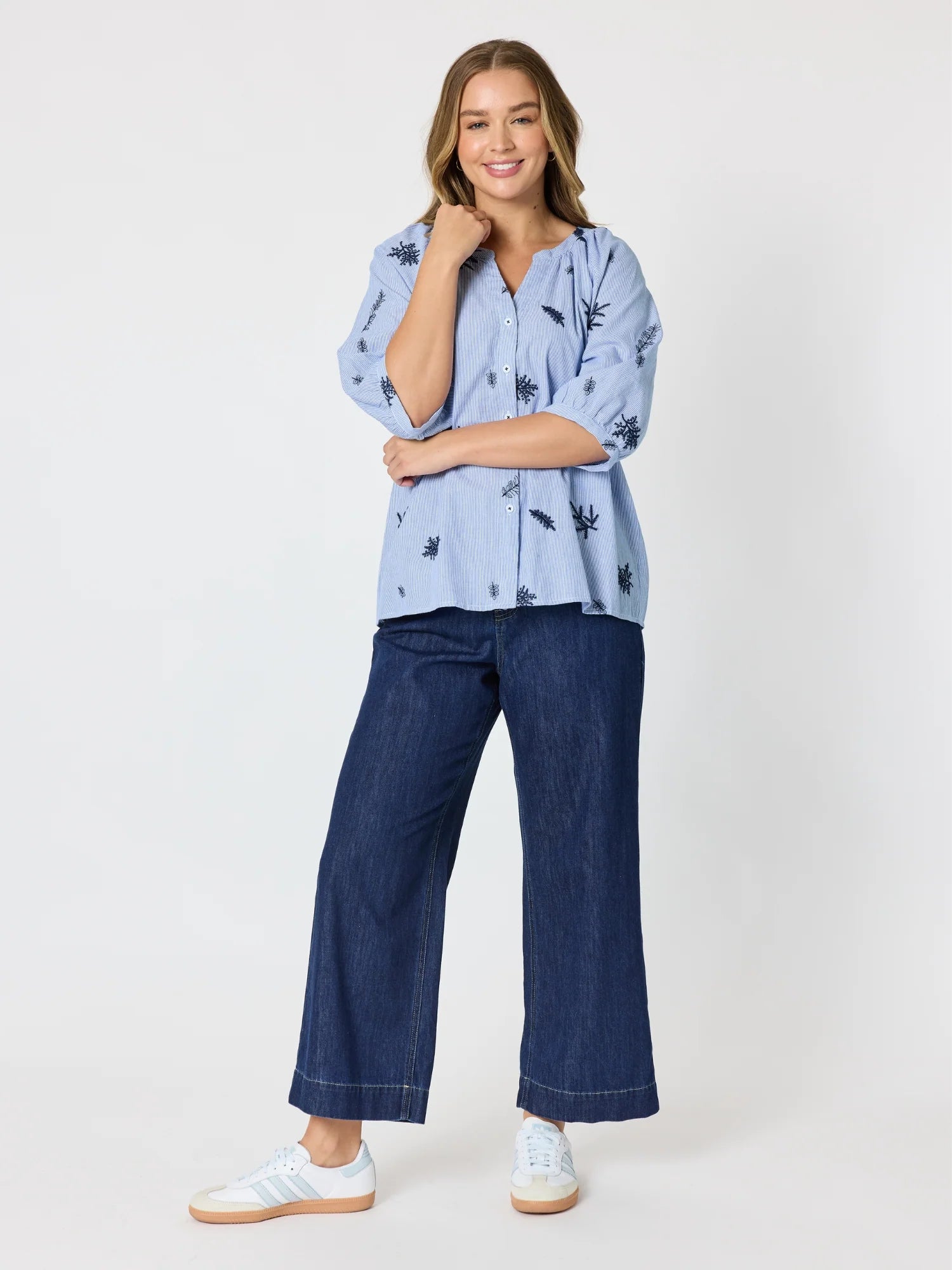 Image of A woman modeling a Threadz light blue short-sleeve button-up shirt with a black leaf pattern, paired with dark blue wide-leg jeans and white sneakers, against a plain white studio background.