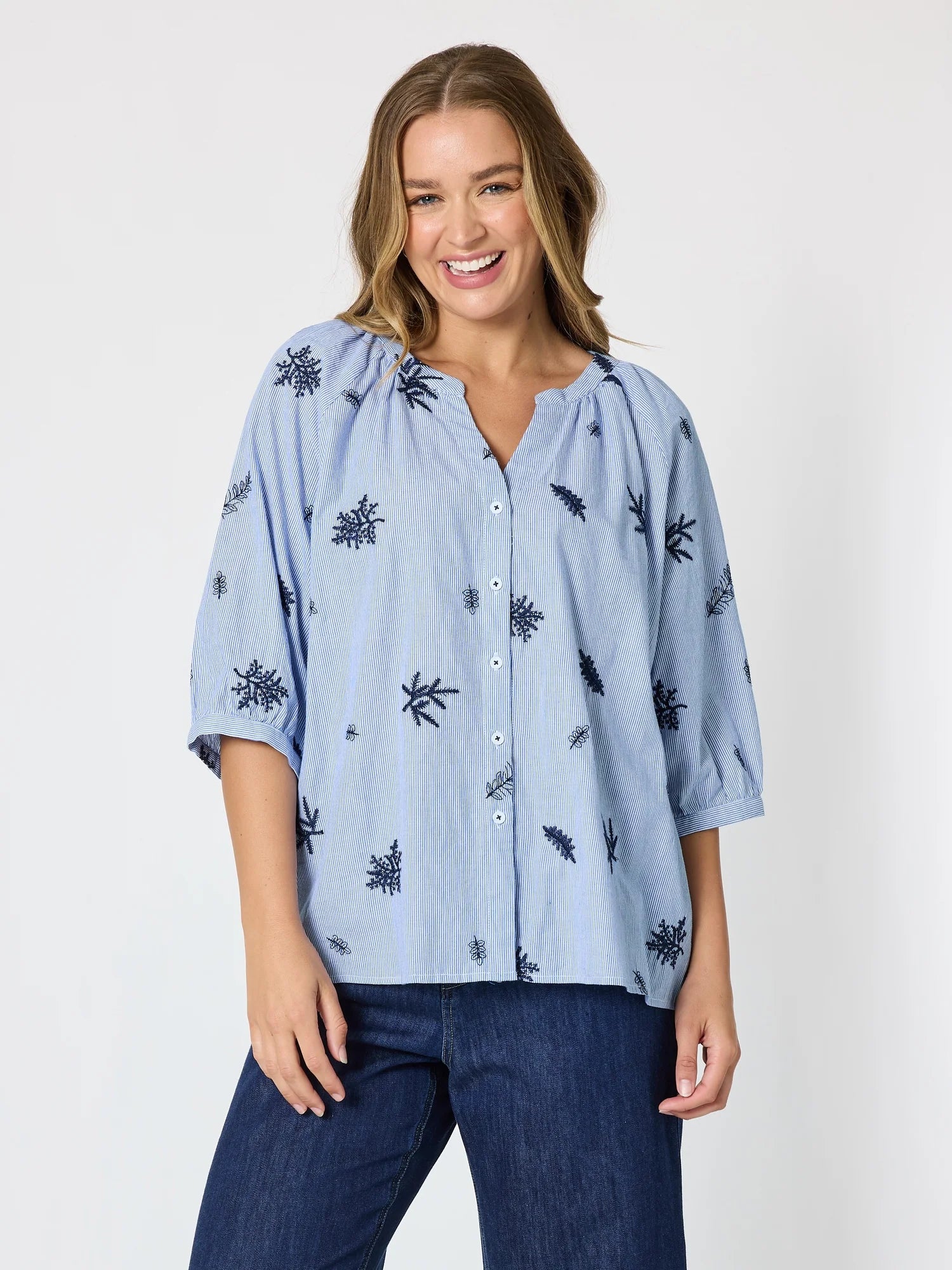 Image of A woman wearing a Threadz light blue button-up blouse with a dark blue leaf pattern and dark blue jeans, standing against a plain white studio background.