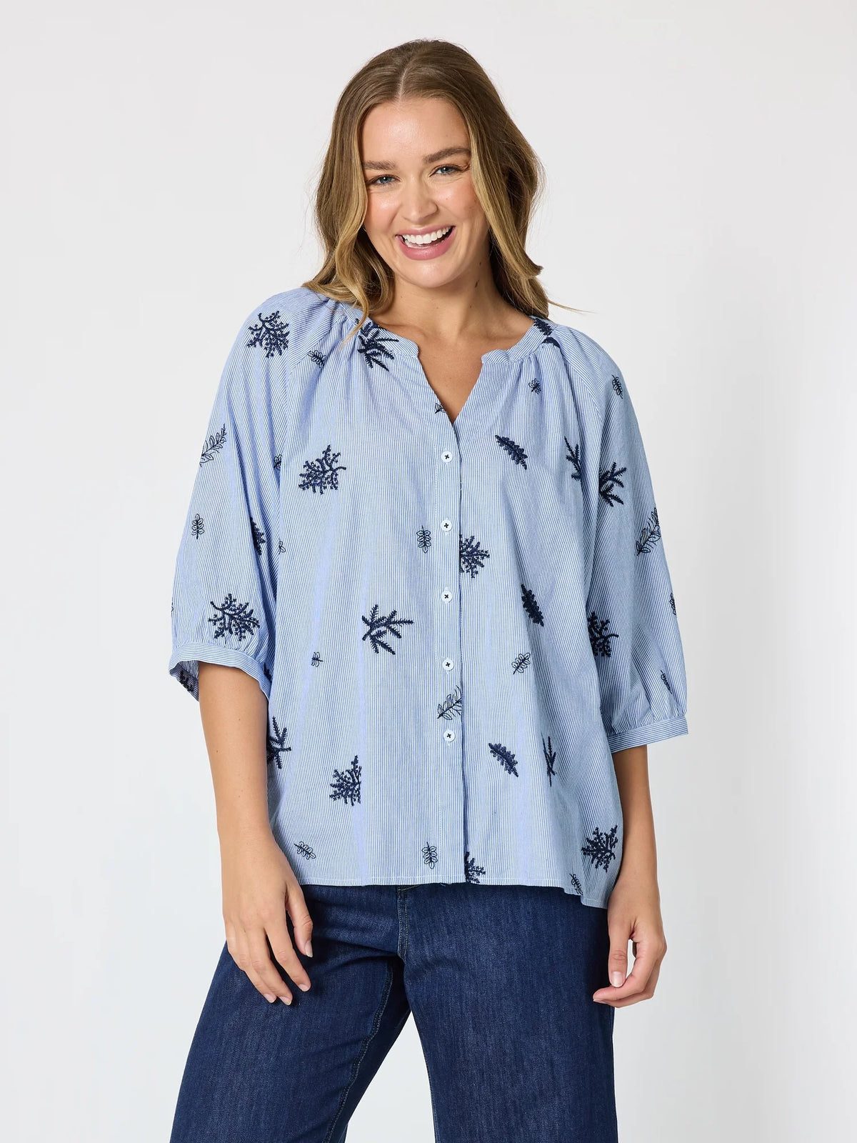 A woman wearing a Threadz light blue button-up blouse with a dark blue leaf pattern and dark blue jeans, standing against a plain white studio background.