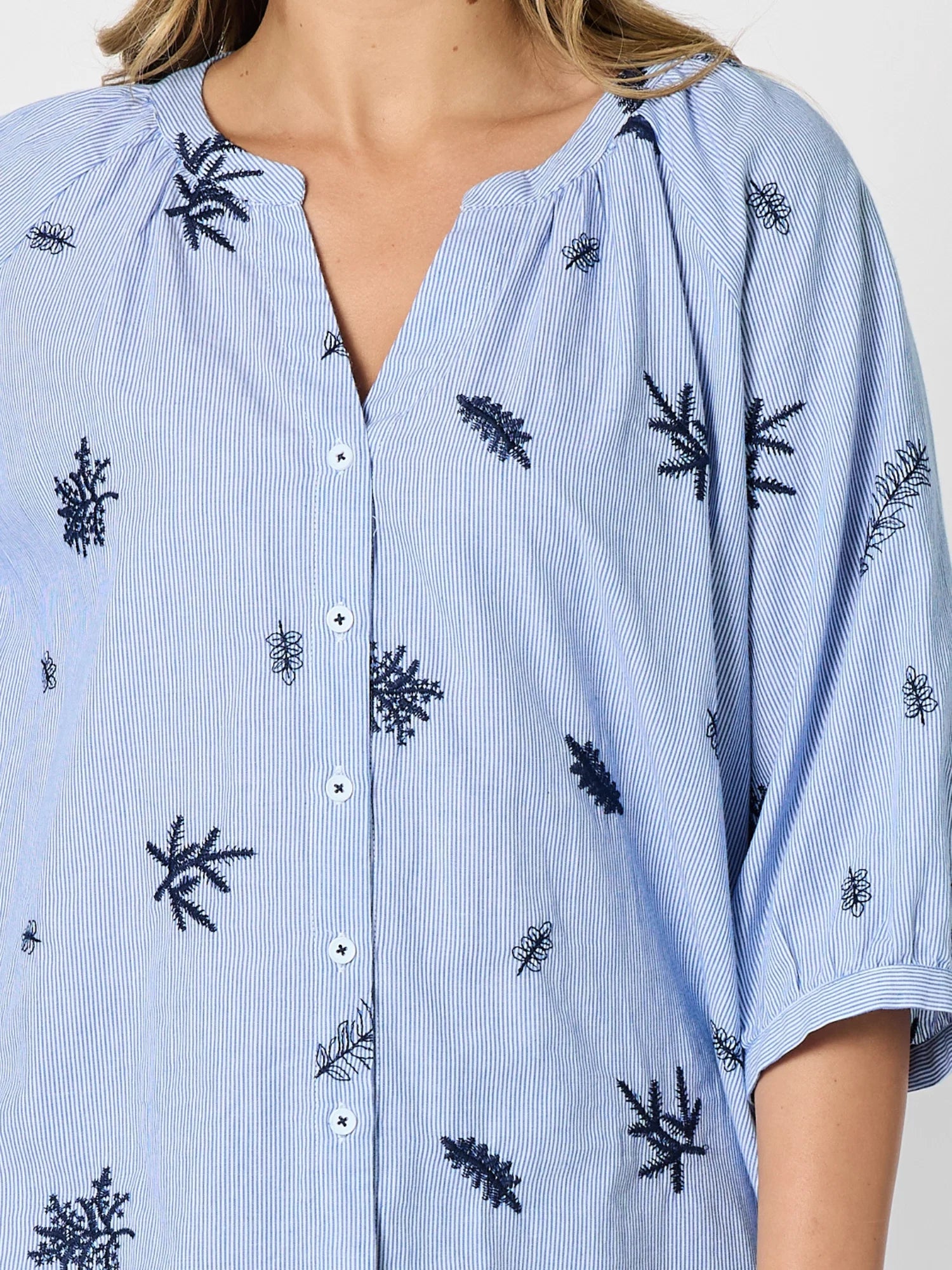 Image of A close-up of a Threadz light blue and white vertically striped button-up shirt with embroidered palm tree motifs, worn by a woman