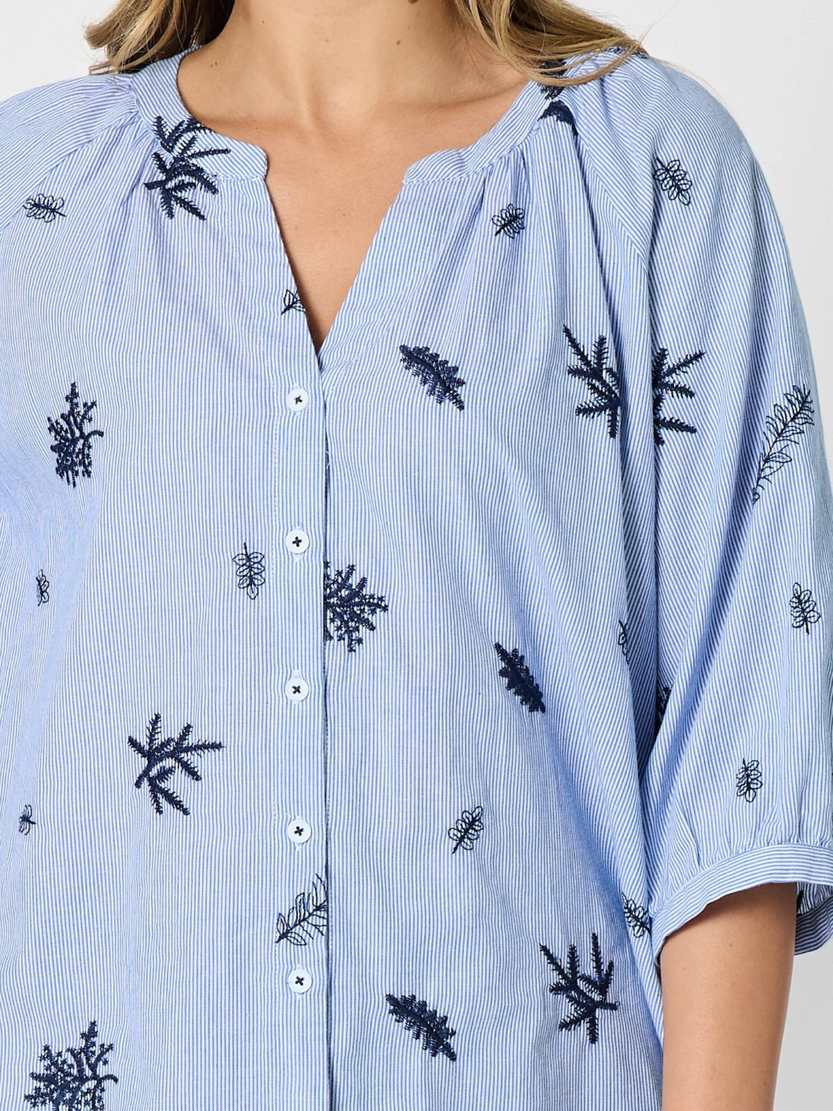 A close-up of a Threadz light blue and white vertically striped button-up shirt with embroidered palm tree motifs, worn by a woman