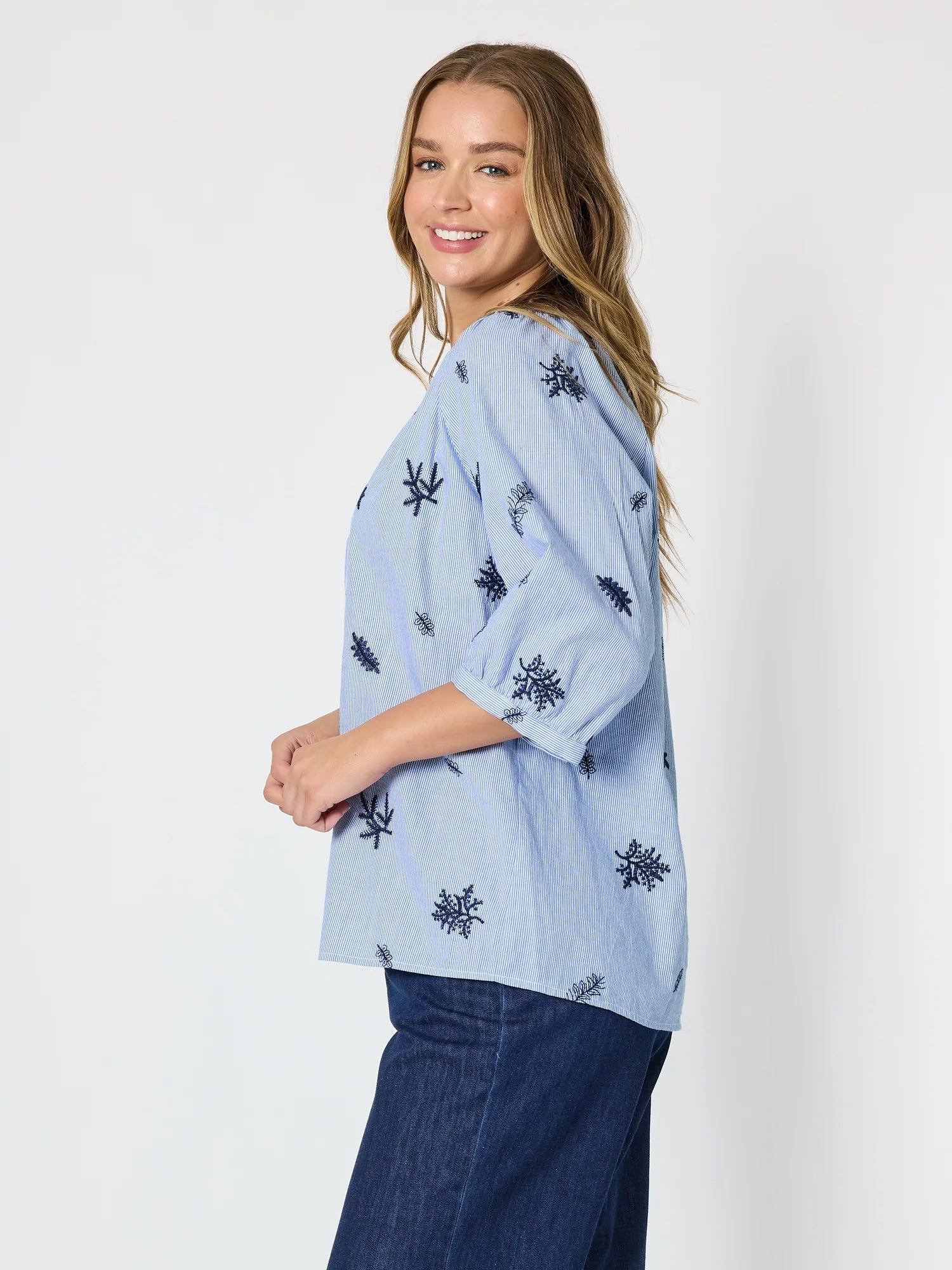 Image of A woman modeling a Threadz light blue cotton blouse with dark navy embroidered snowflake patterns, paired with dark blue jeans, against a plain white studio background.