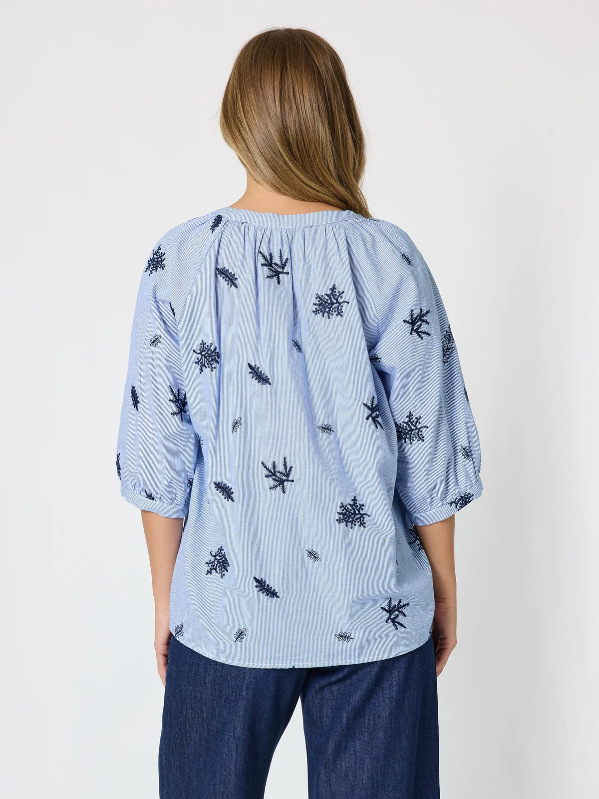 A woman viewed from the back wearing a Threadz light blue blouse with dark navy embroidered leaf motifs and dark blue jeans, set against a plain light gray studio background.