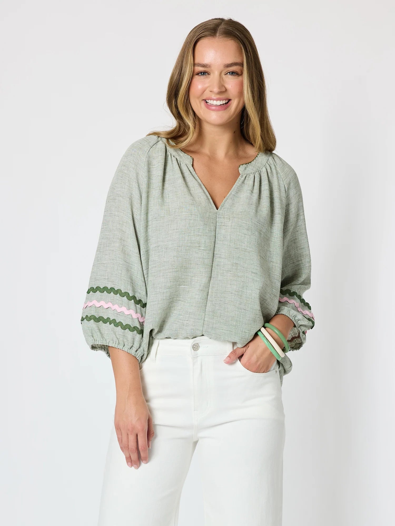 Image of A woman modeling a Threadz sage green long-sleeve blouse with a split neckline and colorful zigzag embroidered trim at the cuffs, paired with white pants against a light gray background.