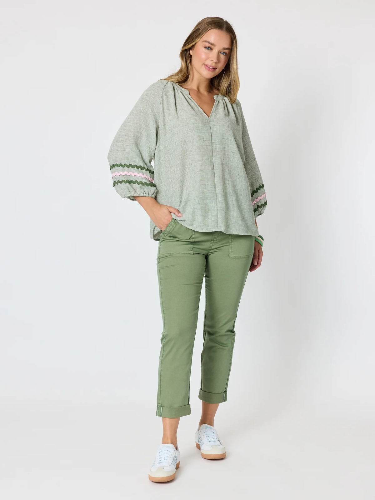 A woman modeling a Threadz light sage green long-sleeve blouse with patterned cuffs and olive green cropped pants, standing against a light gray studio background.