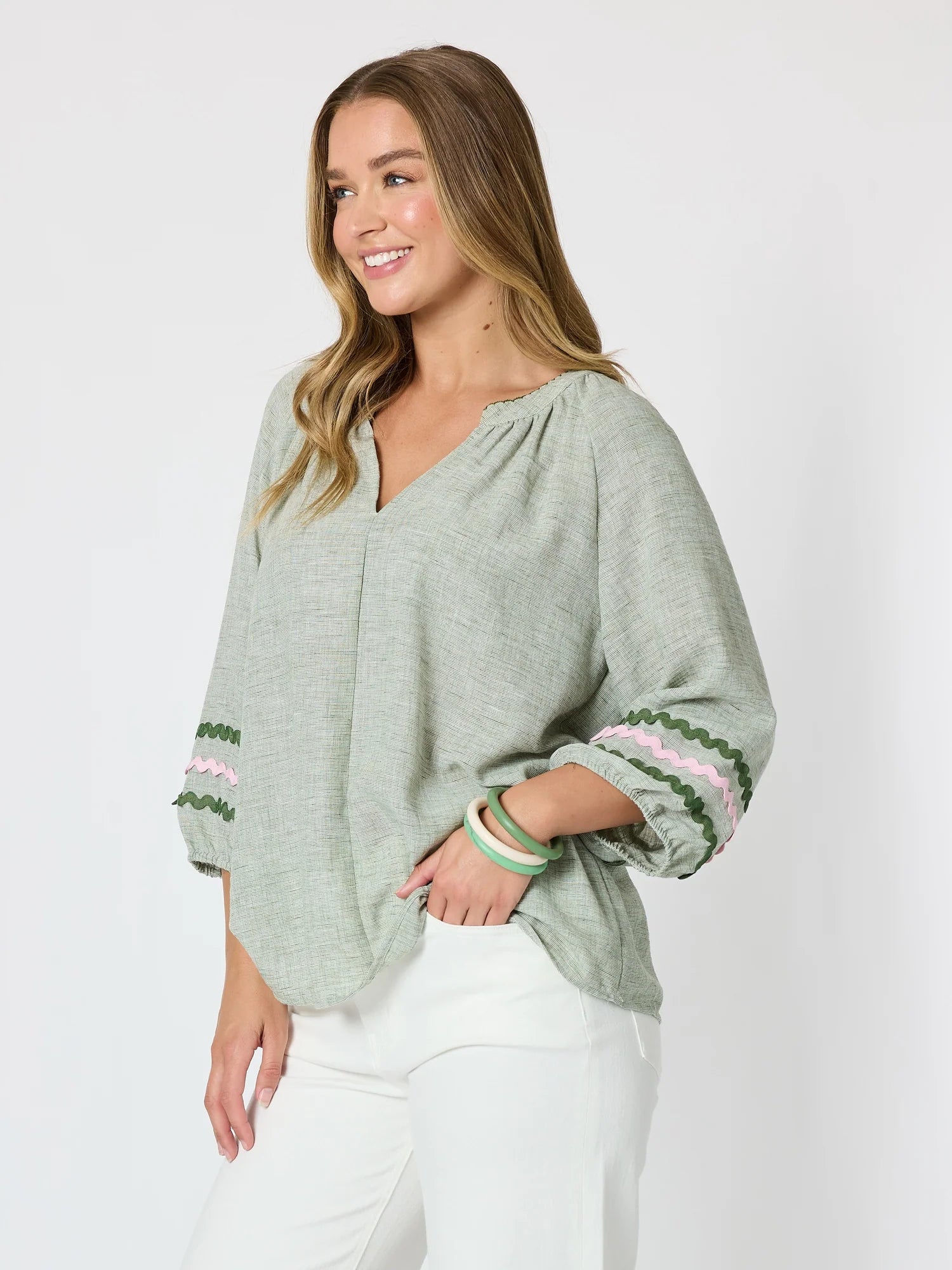 Image of A woman wearing a Threadz light sage green long-sleeve blouse with green and pink embroidered cuffs, paired with white pants, against a plain light gray studio background.