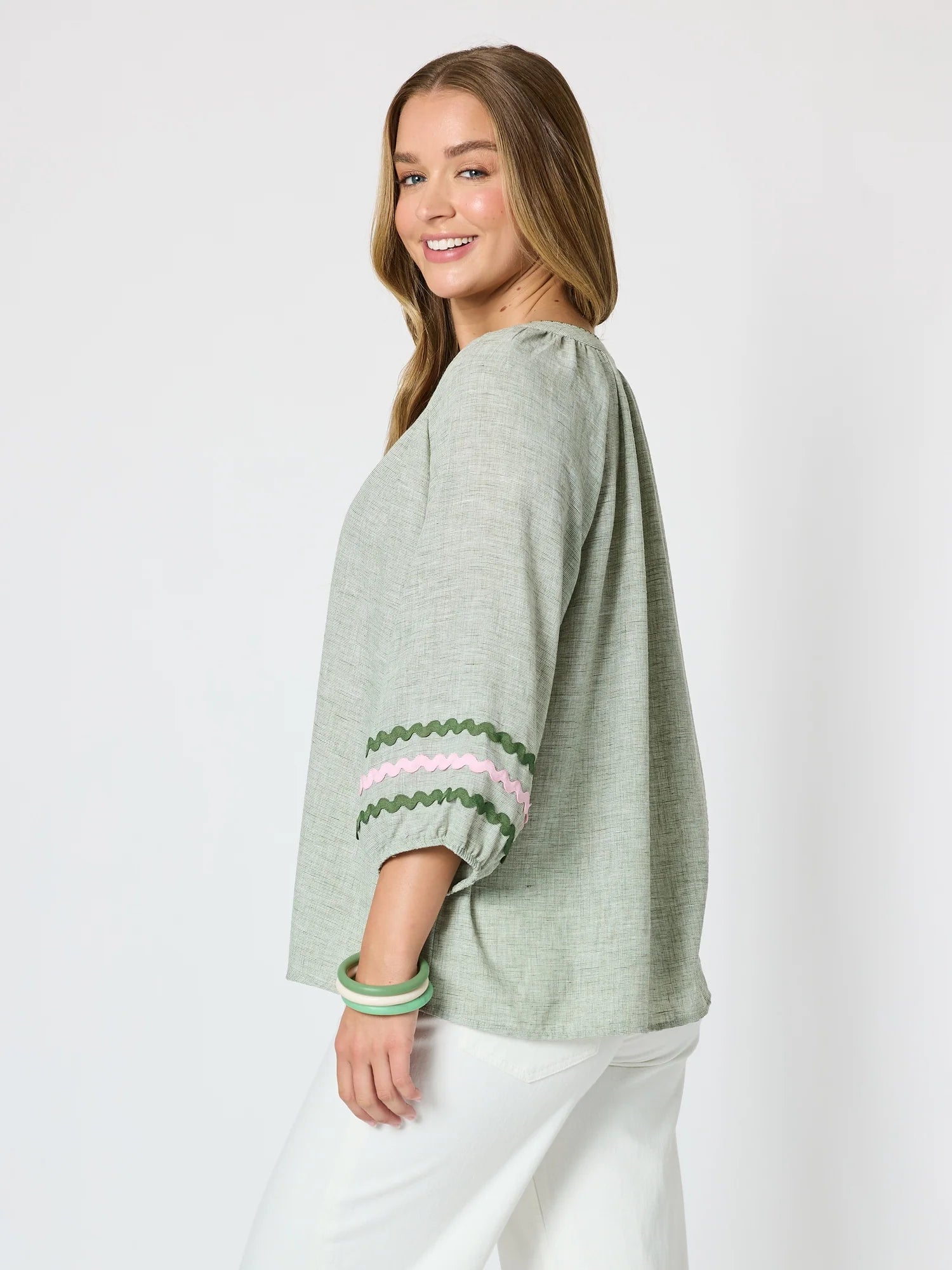 Image of A woman wearing a Threadz sage green linen-blend top with three-quarter sleeves and decorative green, pink, and white striped cuffs, paired with white pants, photographed against a light gray studio background.