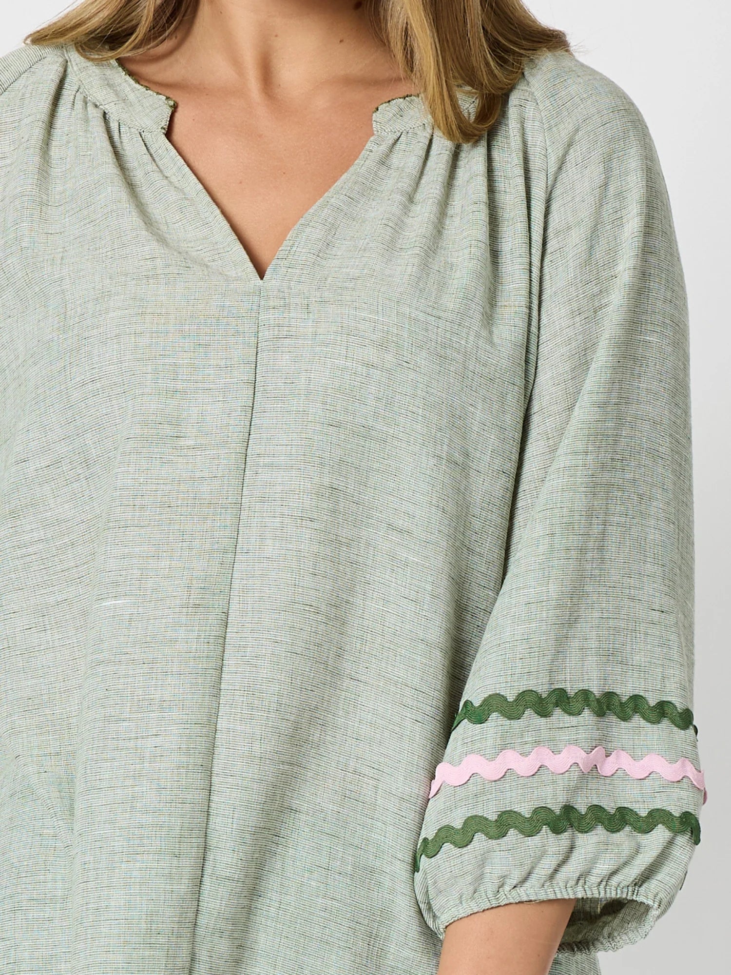 Image of A close-up of a woman wearing a Threadz light sage green linen blouse with three-quarter sleeves. The sleeves feature decorative wavy trim in dark green and light pink near the cuffs.