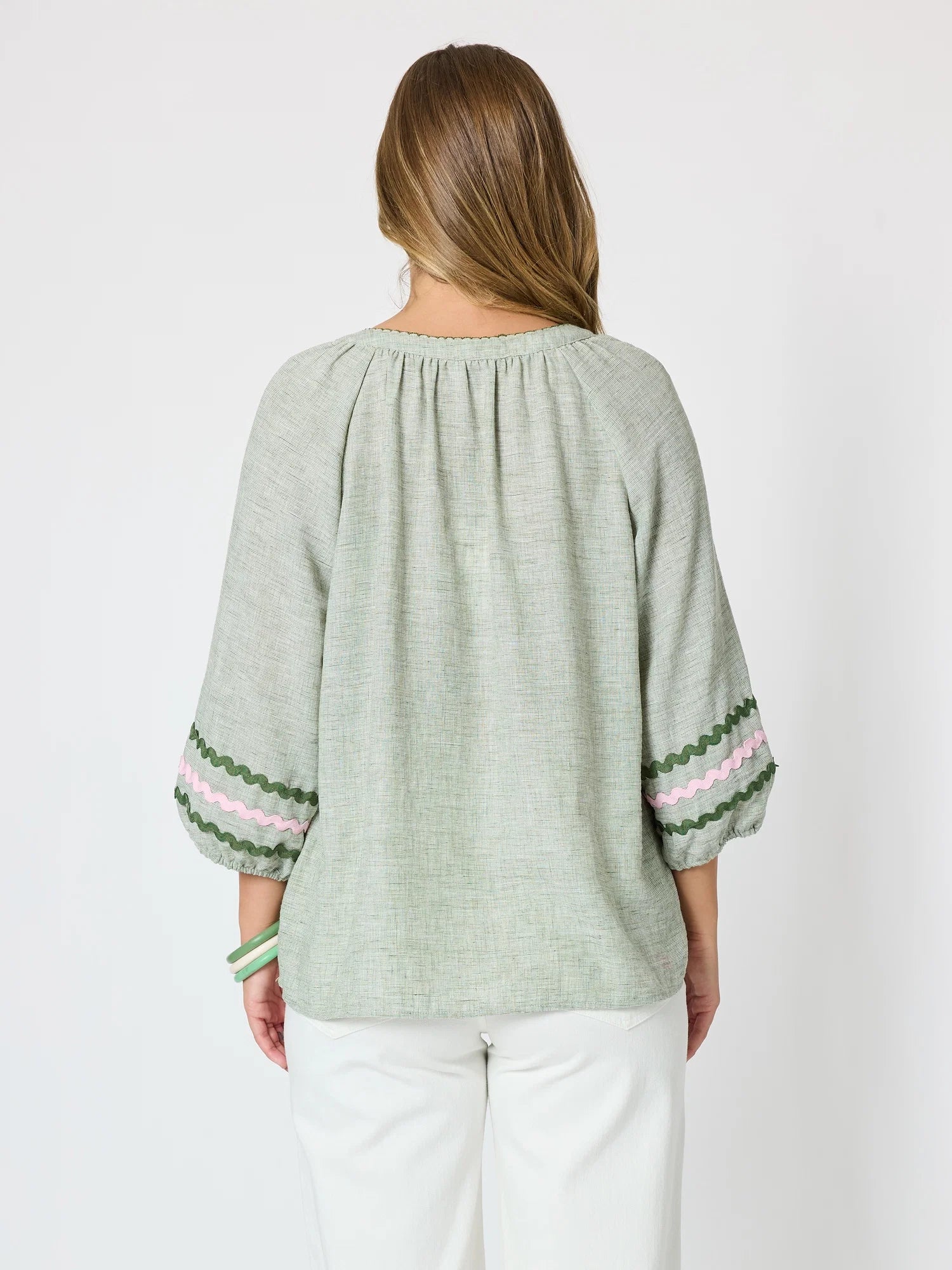 Image of A woman shown from the back wearing a Threadz light sage green linen blouse with three-quarter sleeves and pink and green striped cuffs, paired with white pants, against a plain white background.
