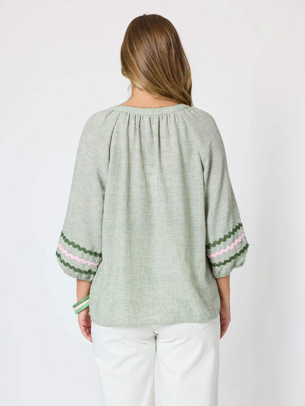 A woman shown from the back wearing a Threadz light sage green linen blouse with three-quarter sleeves and pink and green striped cuffs, paired with white pants, against a plain white background.