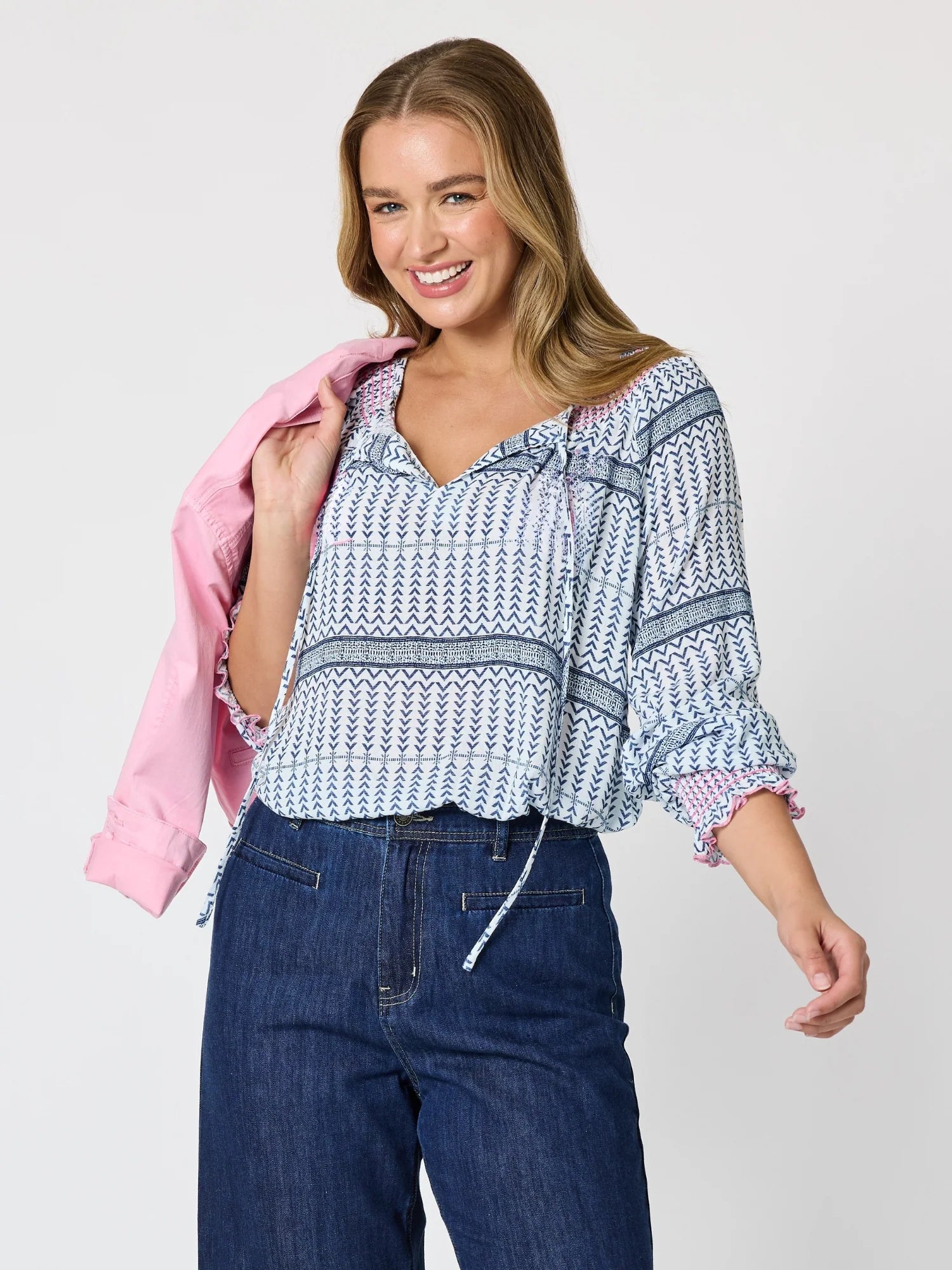 Image of A woman modeling a Threadz blue and white patterned blouse with ruffled sleeves, paired with dark blue jeans and holding a light pink jacket against a light gray studio background.