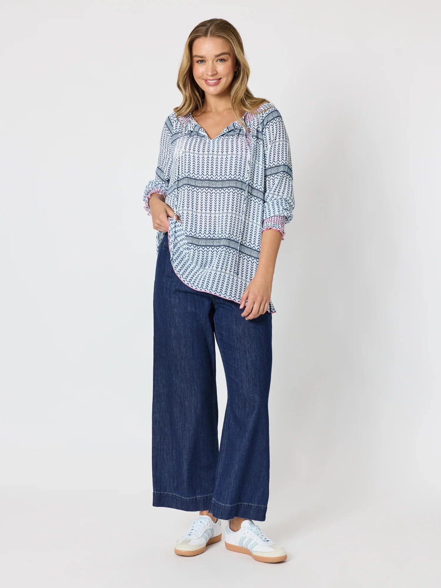 Image of A woman modeling a Threadz loose, long-sleeve button-up shirt with a blue and white geometric pattern, paired with wide-leg dark blue jeans and white sneakers, against a plain light gray studio background.