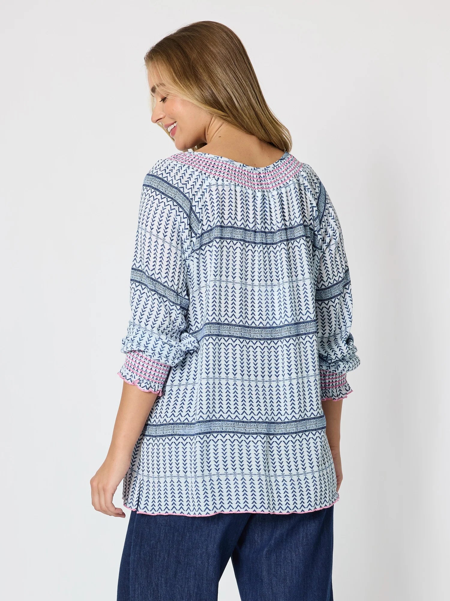 Image of A woman shown from the back wearing a Threadz long-sleeve patterned blouse with blue, white, and pink geometric stripes, paired with dark blue jeans, against a light gray studio background.