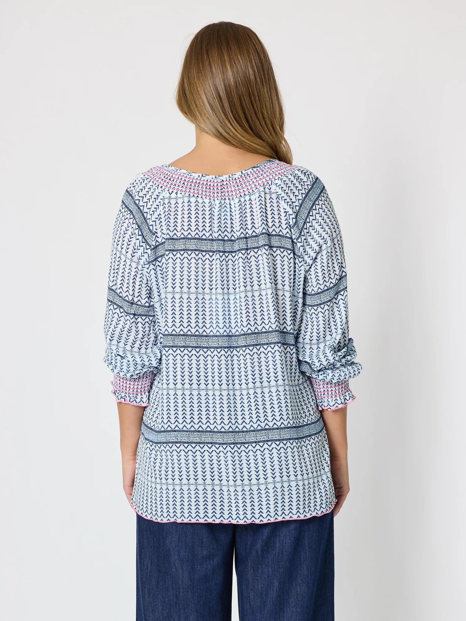 Image of A woman viewed from the back wearing a Threadz patterned long-sleeve top with blue, white, and pink geometric stripes, paired with dark blue jeans, against a plain light gray background.
