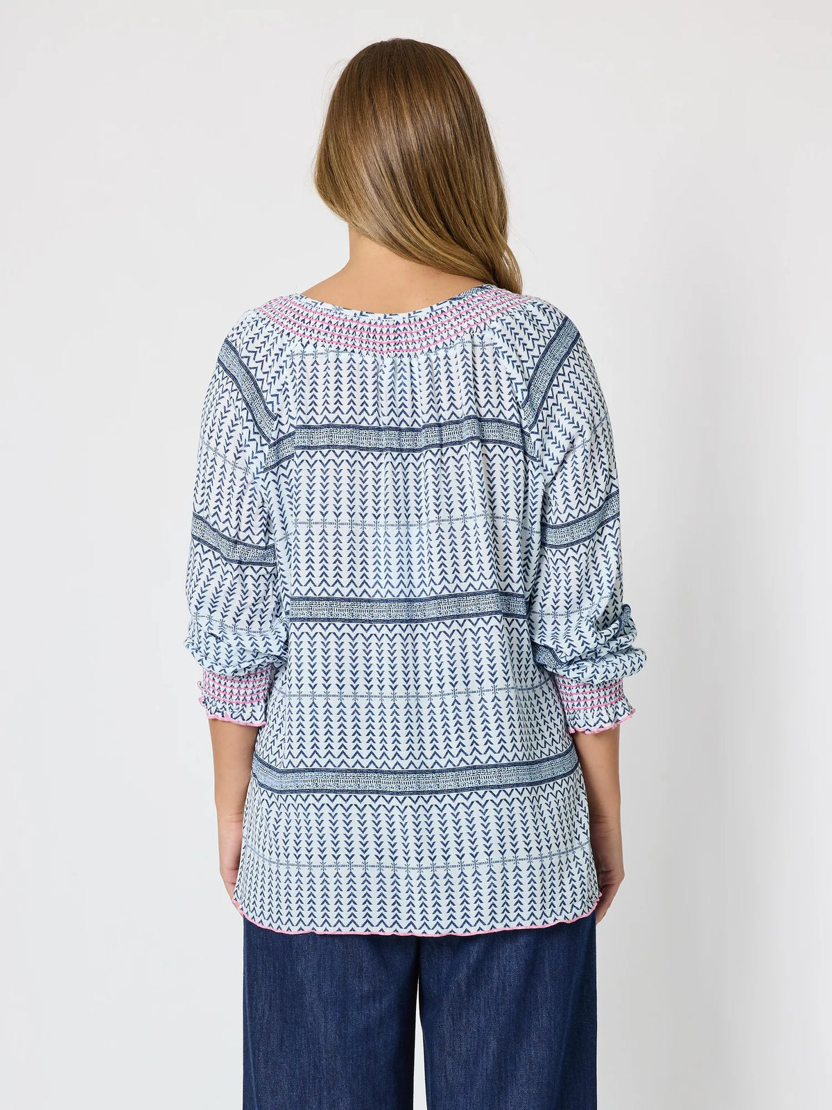 A woman viewed from the back wearing a Threadz patterned long-sleeve top with blue, white, and pink geometric stripes, paired with dark blue jeans, against a plain light gray background.
