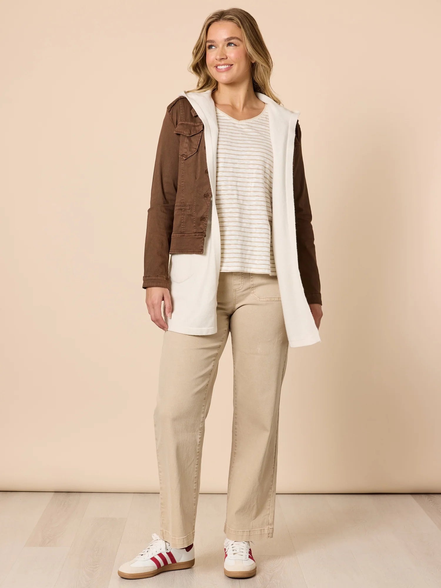 Woman wearing a brown jacket, white shirt, Threadz beige pants, and white sneakers on a beige background