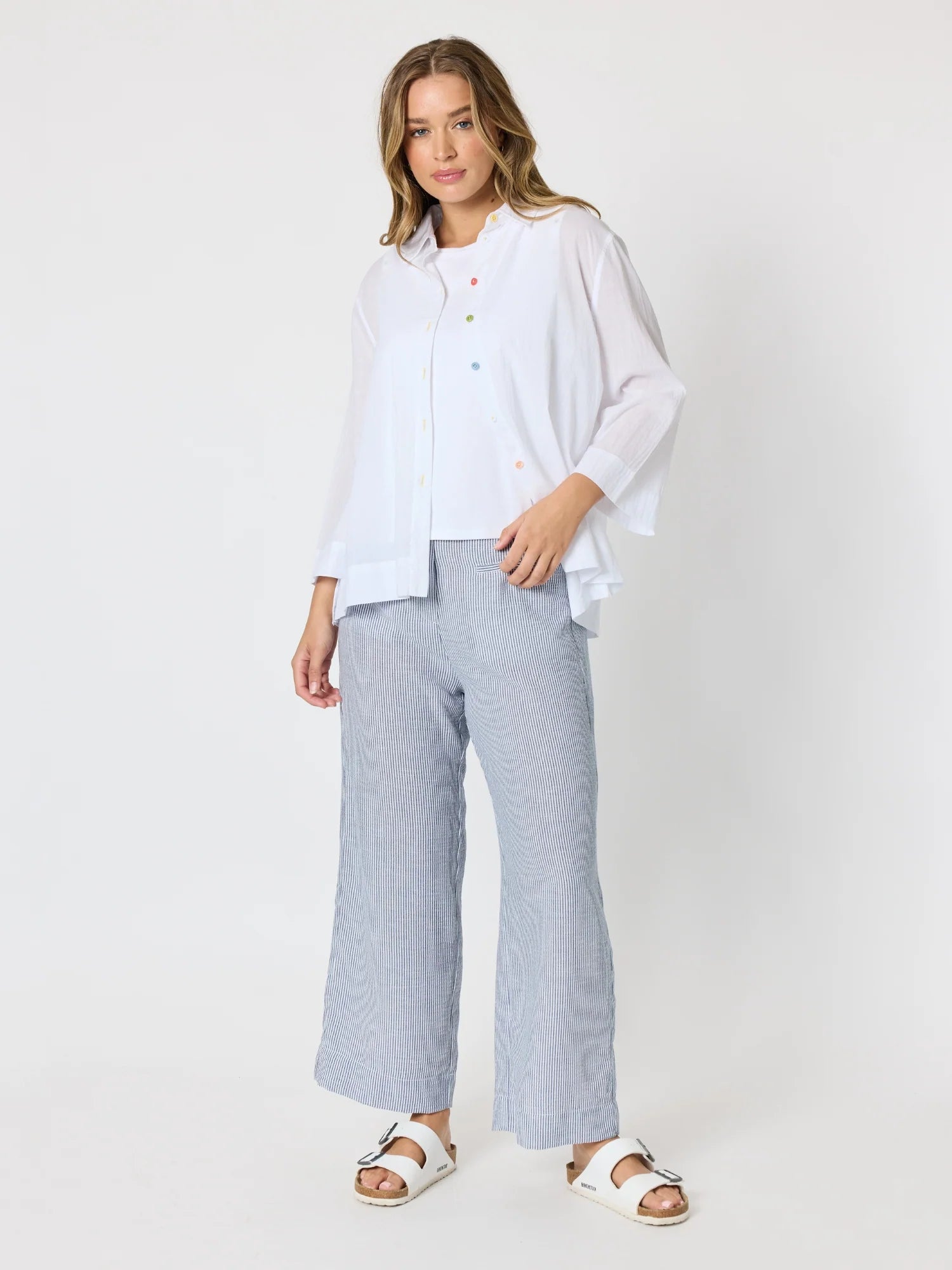 Image of A woman modeling a white long-sleeve button-up blouse and Threadz light blue striped wide-leg trousers, paired with white slide sandals, against a plain white studio background.