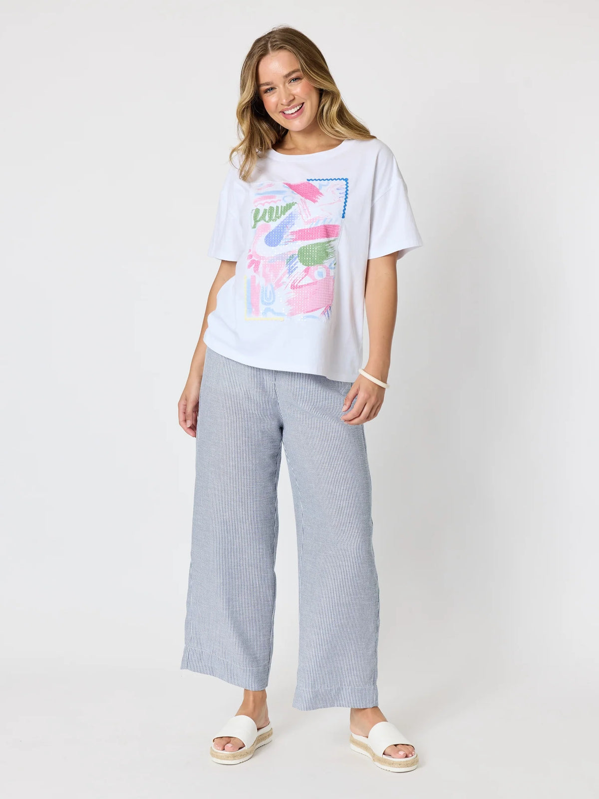 A woman modeling a white graphic t-shirt with a colorful abstract print, paired with Threadz light blue wide-leg cropped pants and white slide sandals, against a plain light gray studio background.