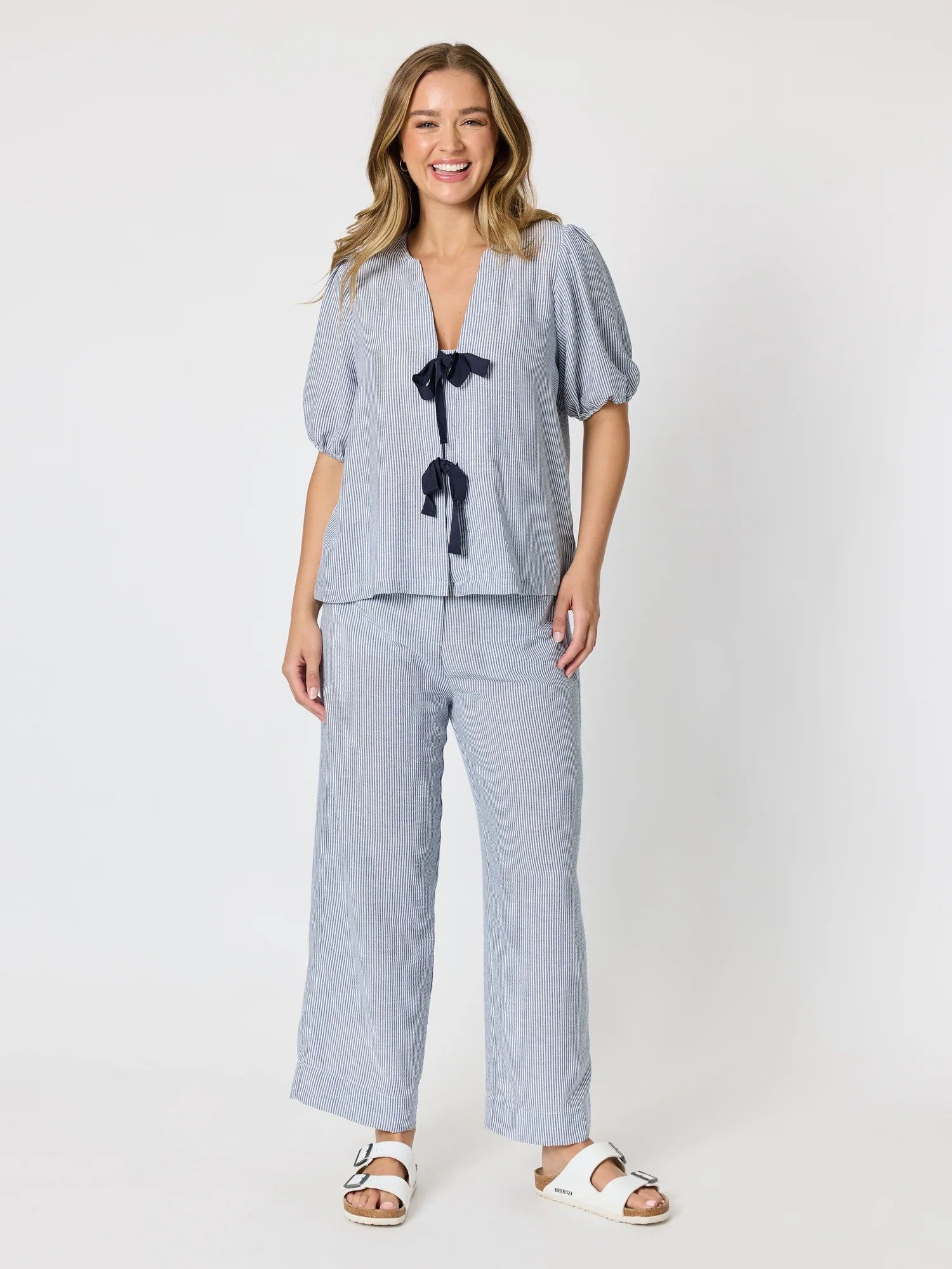 Image of A woman modeling a Threadz light blue linen two-piece set consisting of a short-sleeve blouse with a black bow detail and matching wide-leg trousers, paired with white slide sandals, against a plain white studio background.