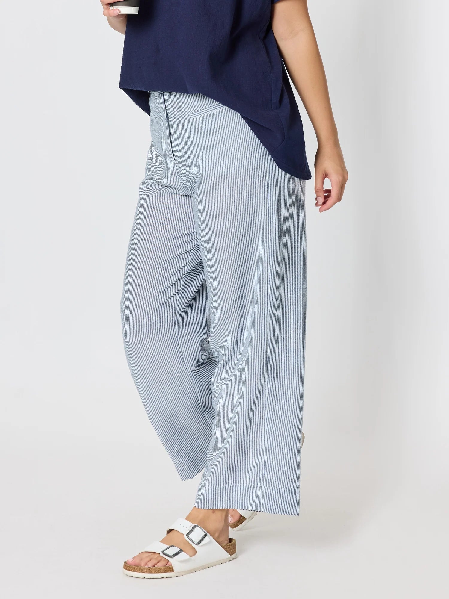 Image of A woman wearing Threadz light blue wide-leg pleated trousers, a navy blue top, and white slide sandals, shown from the waist down against a plain light gray studio background.