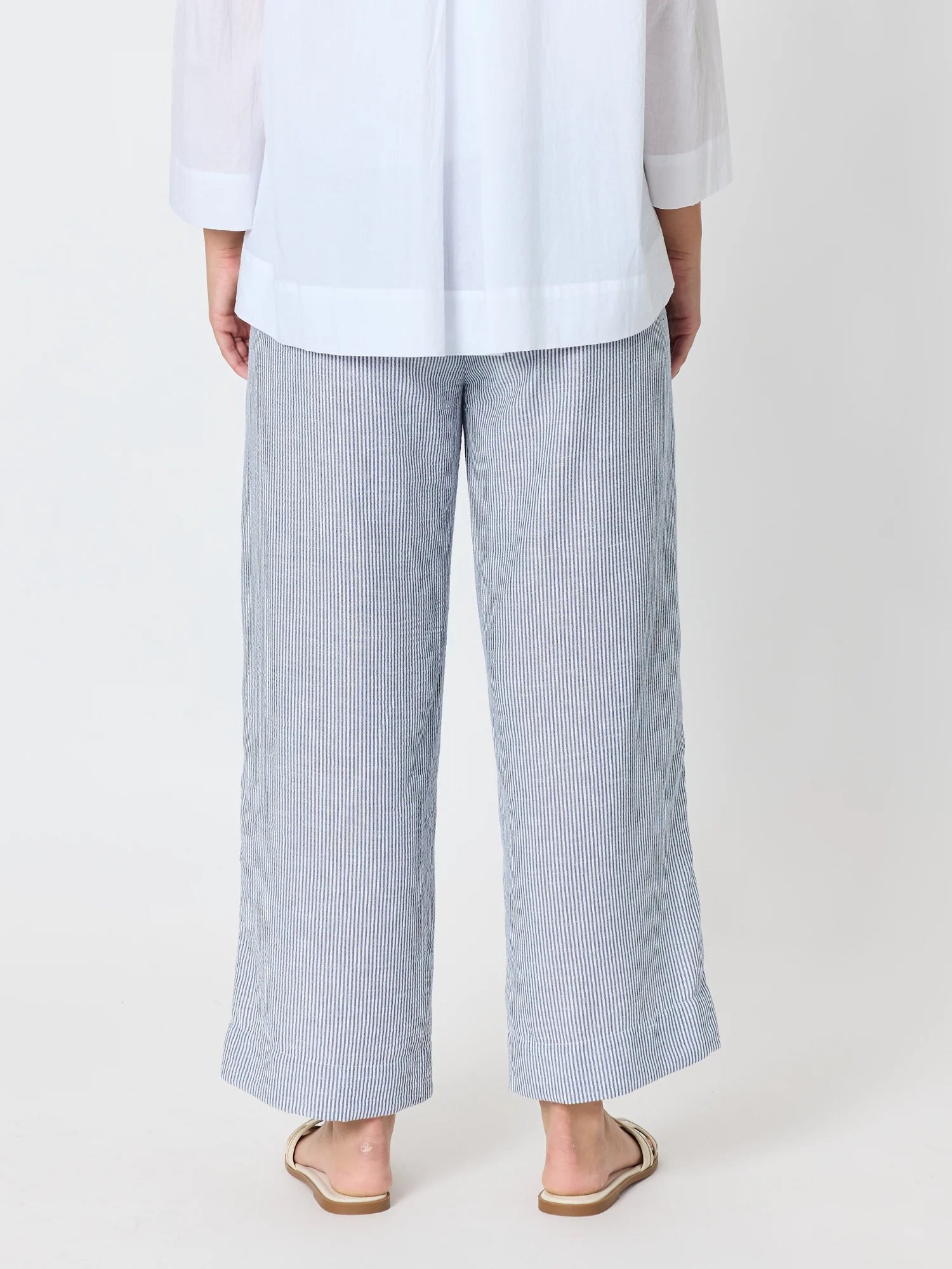 Image of A woman shown from the back wearing Threadz light blue and white vertically striped wide-leg cropped pants, a white blouse, and beige flat sandals against a plain light background.
