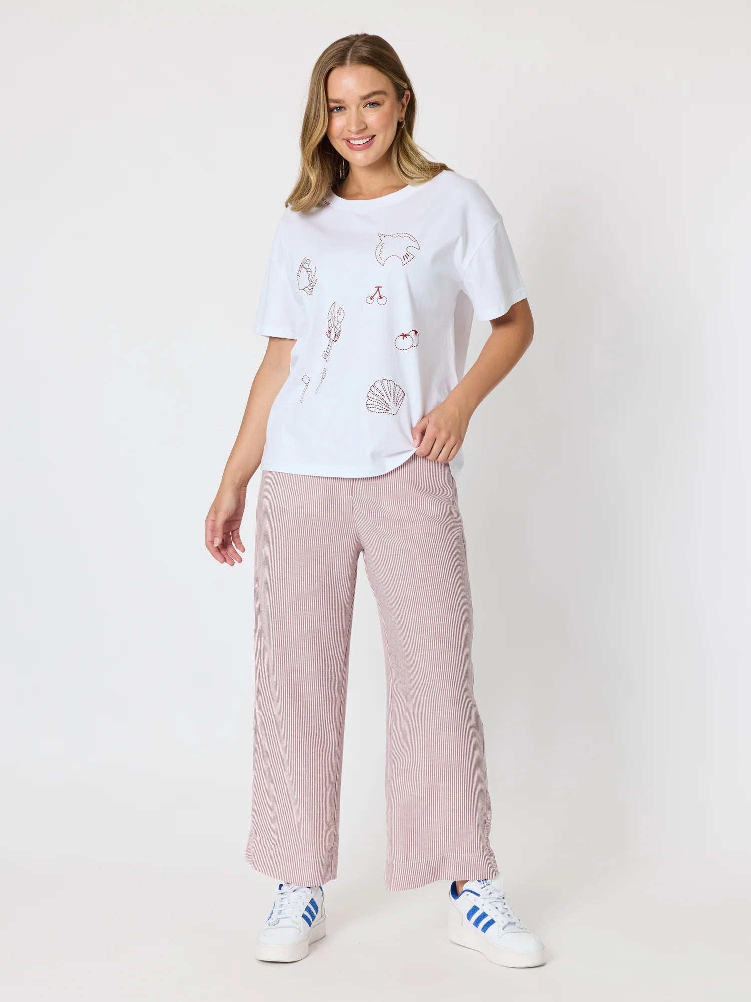 Image of A woman modeling a white short-sleeve t-shirt with delicate pink line-art illustrations and Threadz matching wide-leg dusty pink trousers, paired with white sneakers, against a plain white studio background.
